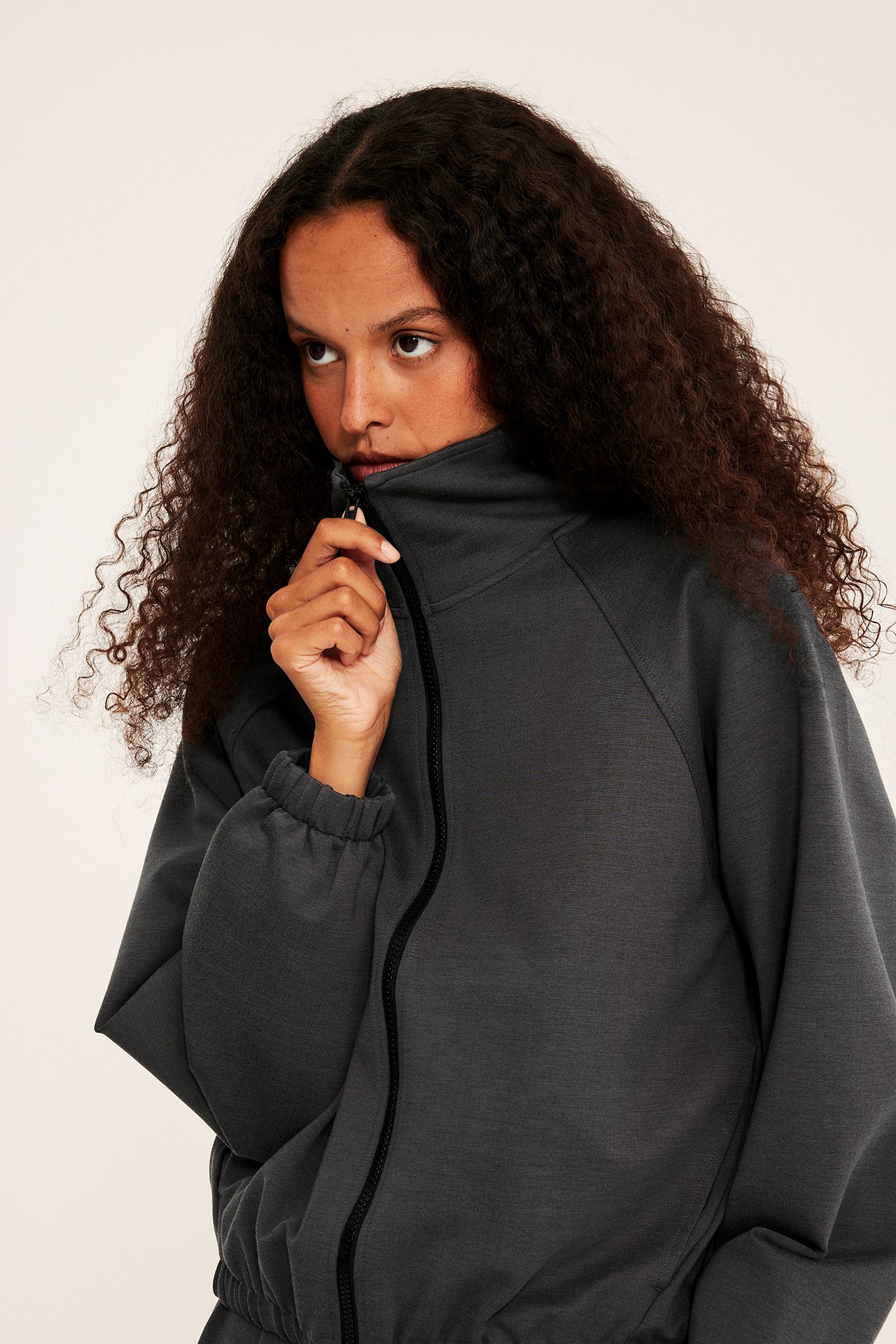 Närbild av kvinnlig modell som drar upp en mörkgrå full-zip sweatshirt med hög krage.