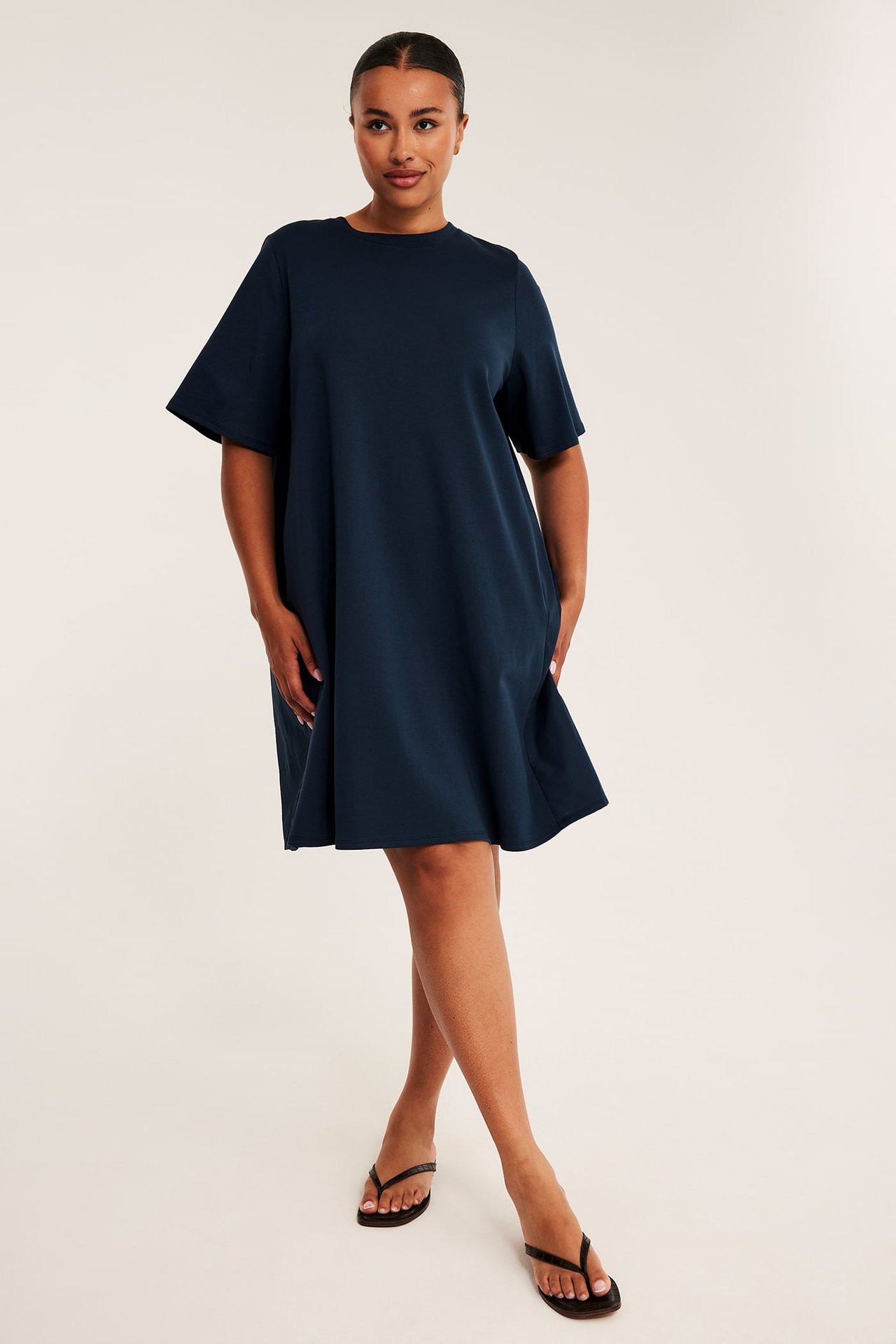 Frontvy av en kvinnlig modell i marinblå oversized t-shirtklänning och svarta flip-flops.