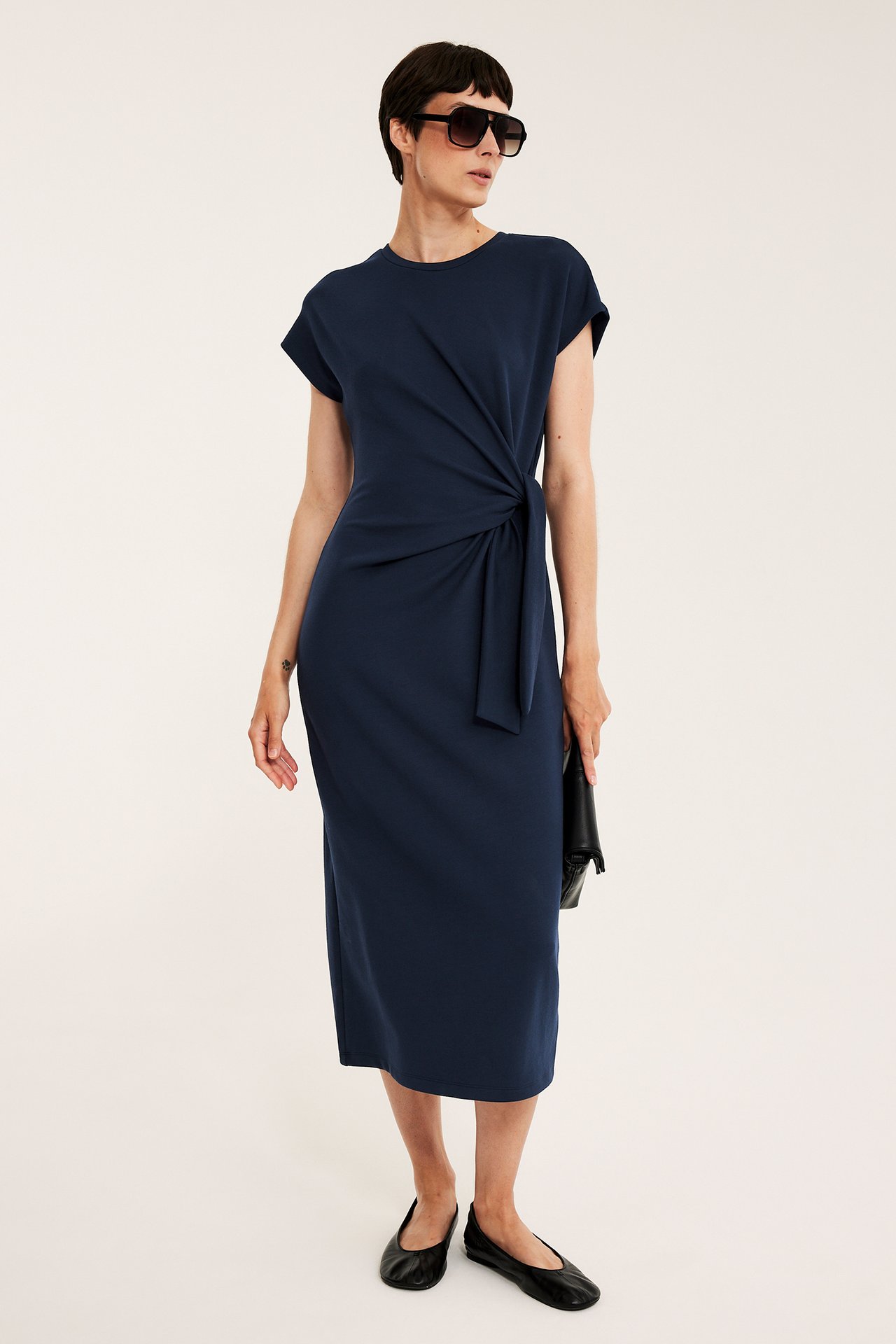 Framifrån, kvinna i mörkblå lång jerseyklänning med sidoknyt, solglasögon, svart clutch och ballerinaskor.