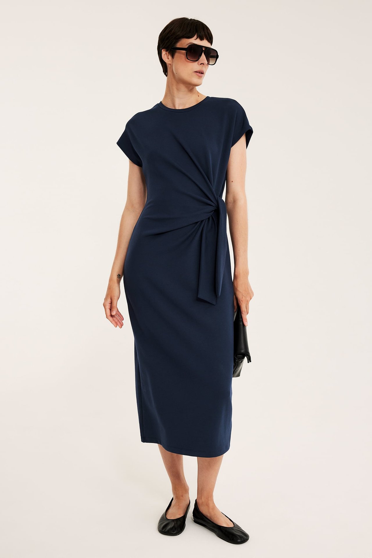 Frontutsikt av kvinne i mørkeblå lang jerseykjole med knyting i siden, solbriller, svart clutch og ballerinasko.