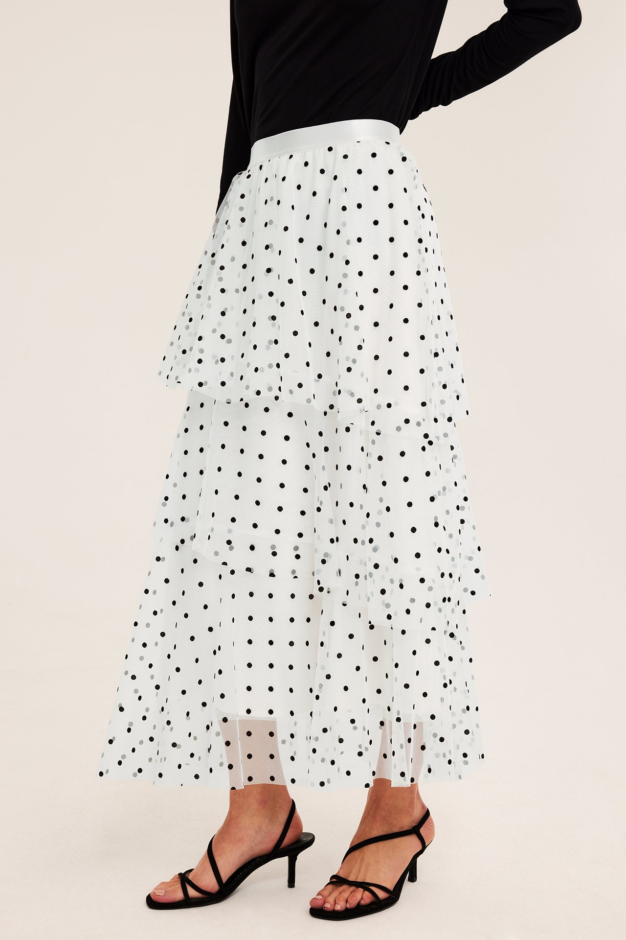 Front view: female model in vit prickig volang midi-kjol, svart långärmad topp och svarta remsandaler.