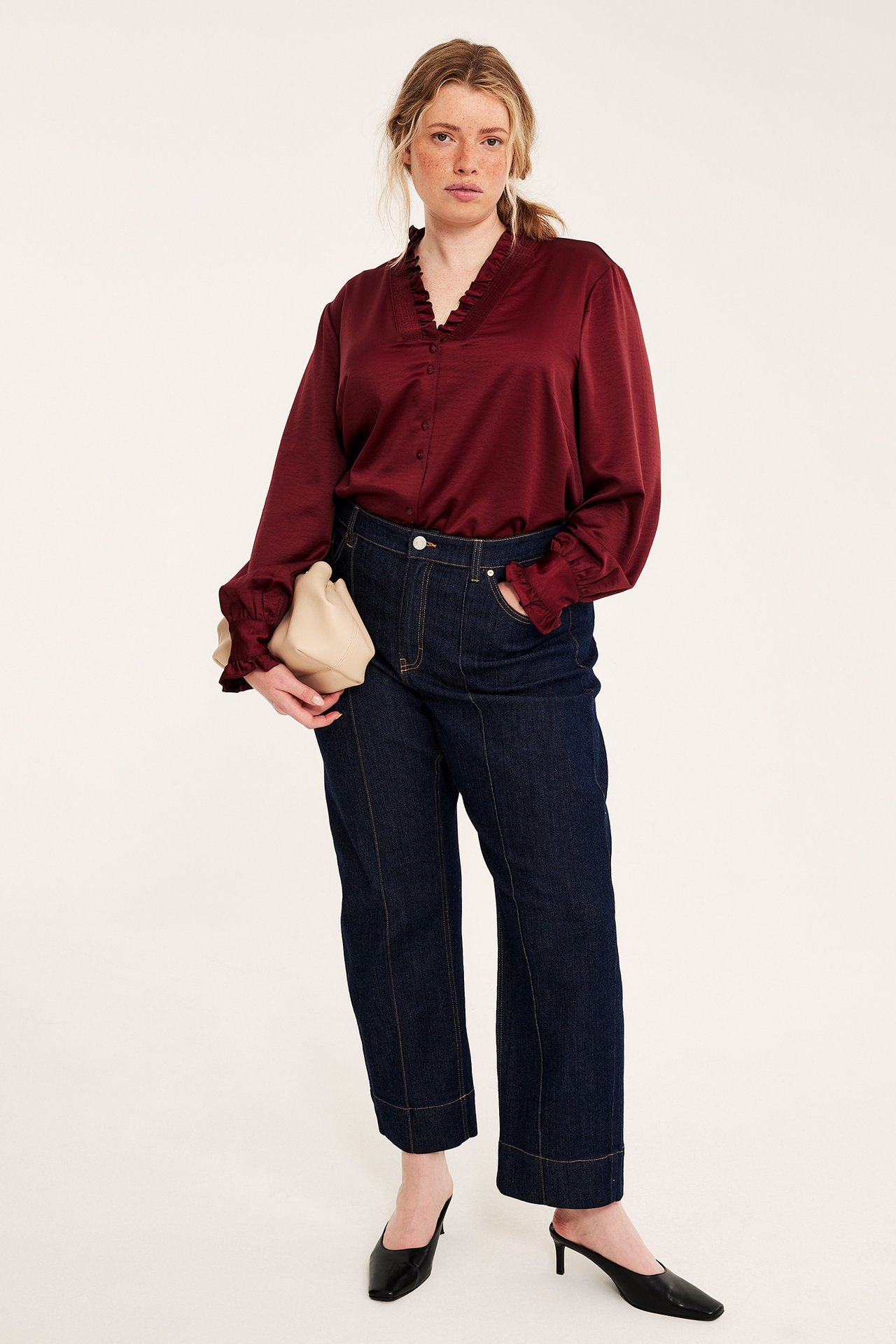 Frontvy av kvinna i rödbrun volangblus, mörkblå jeans, svarta mules, beige clutch.