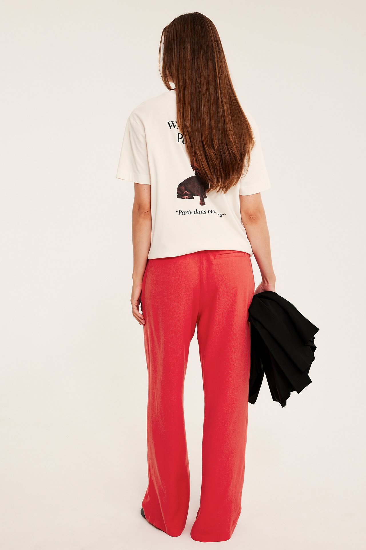Baksidan av kvinna i offwhite grafisk t-shirt, röda vida byxor, håller svart jacka.