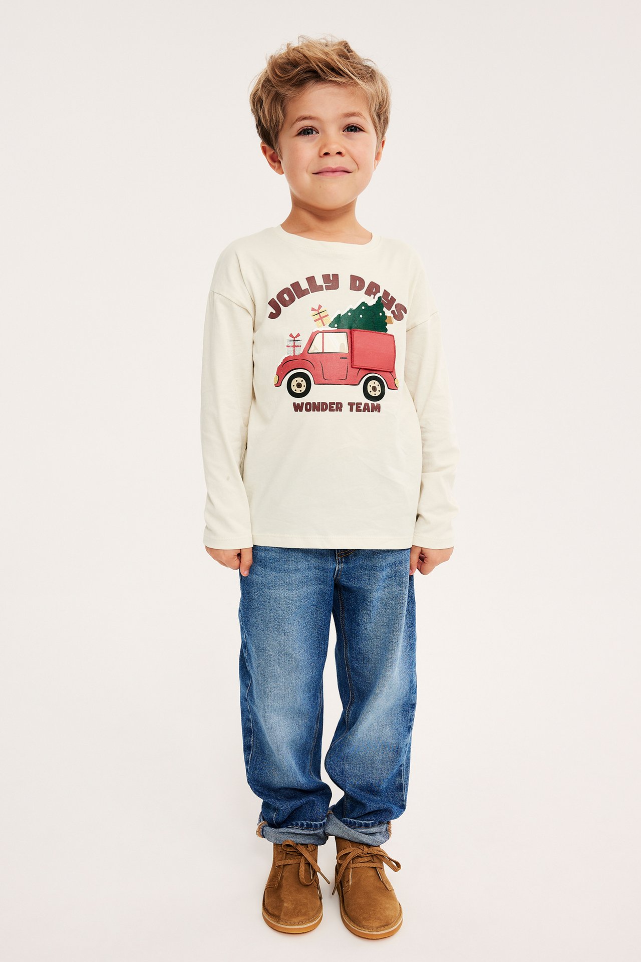 Barn i off-white langermet t-skjorte med juletrykk, blå jeans og brune støvler, forfra.