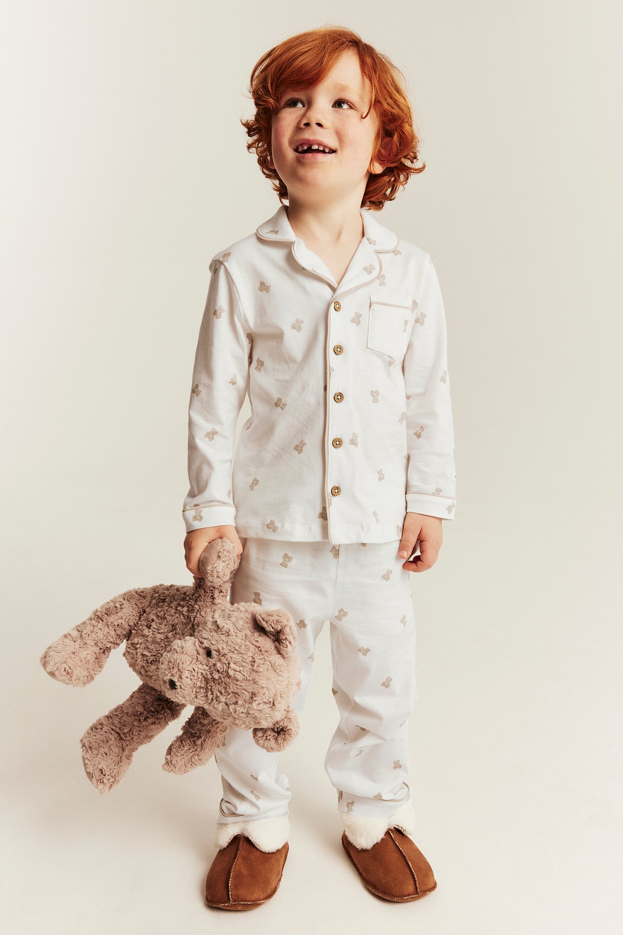 Frontvy av barn i vit pyjamas med nallebjörnsmönster, håller en brun nallebjörn och bruna tofflor.