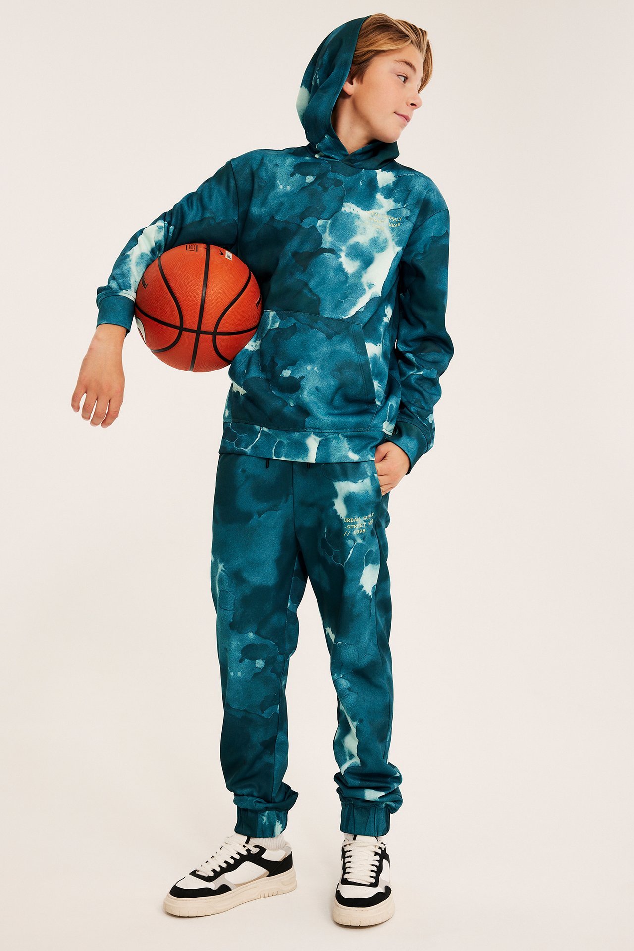 Frontbilde av et barn i turkis tie-dye hettegenser, matchende joggebukse, joggesko, holder basketball.