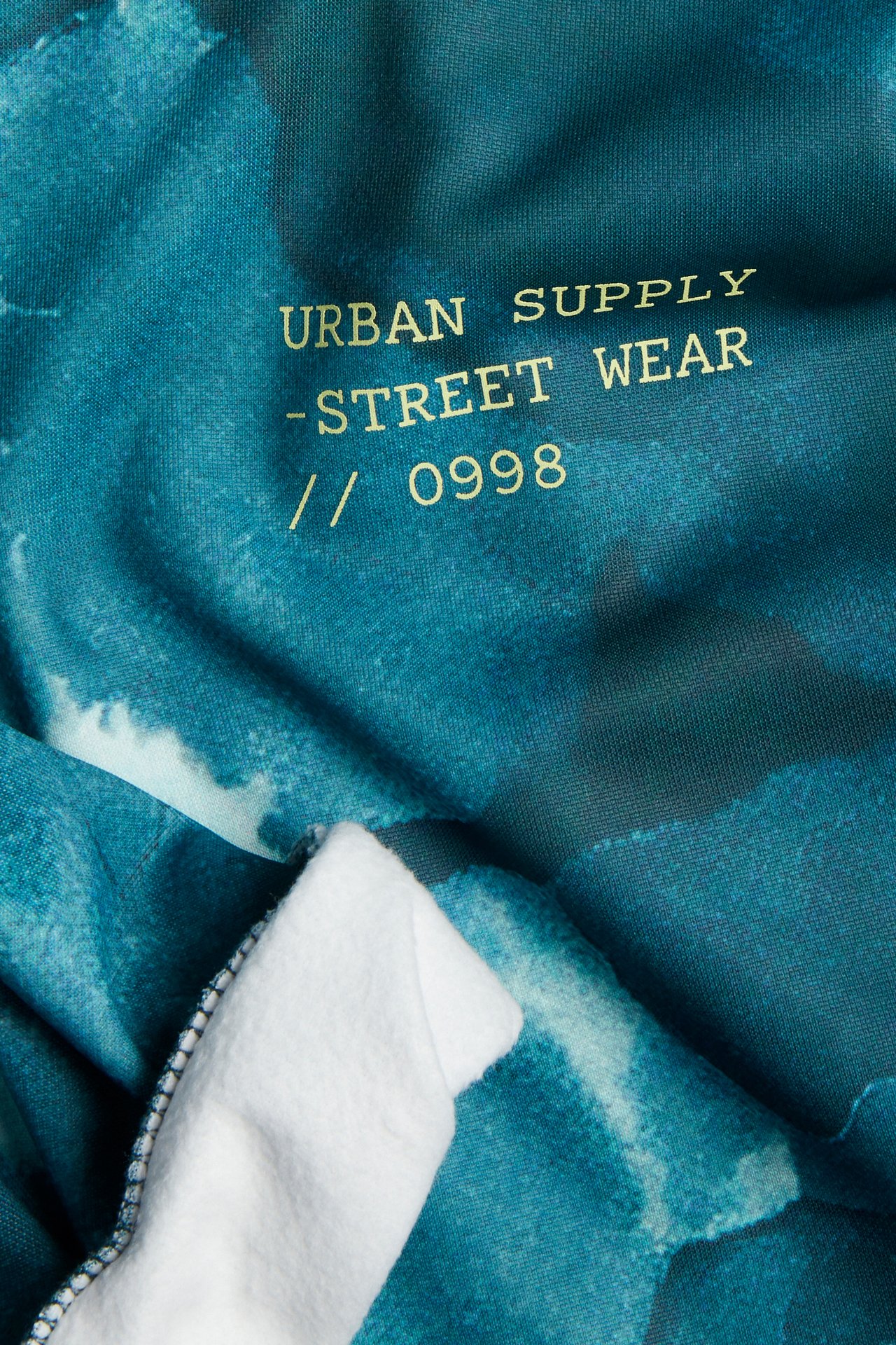 Närbild av ett turkost tyg med guldfärgat 'URBAN SUPPLY - STREET WEAR' tryck och vitt fleecefoder.