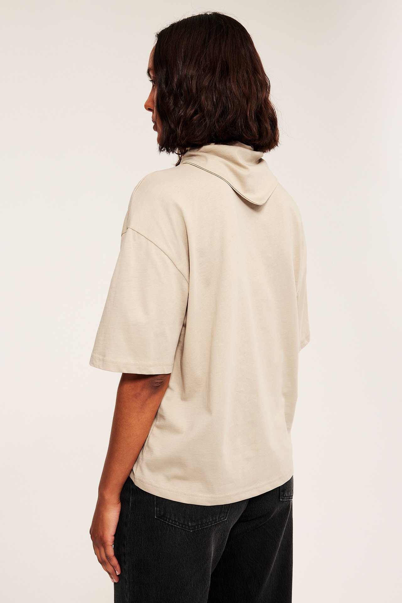 Sidovy bakifrån: oversized ljusbeige kortärmad topp med draperad hals och svarta jeans.