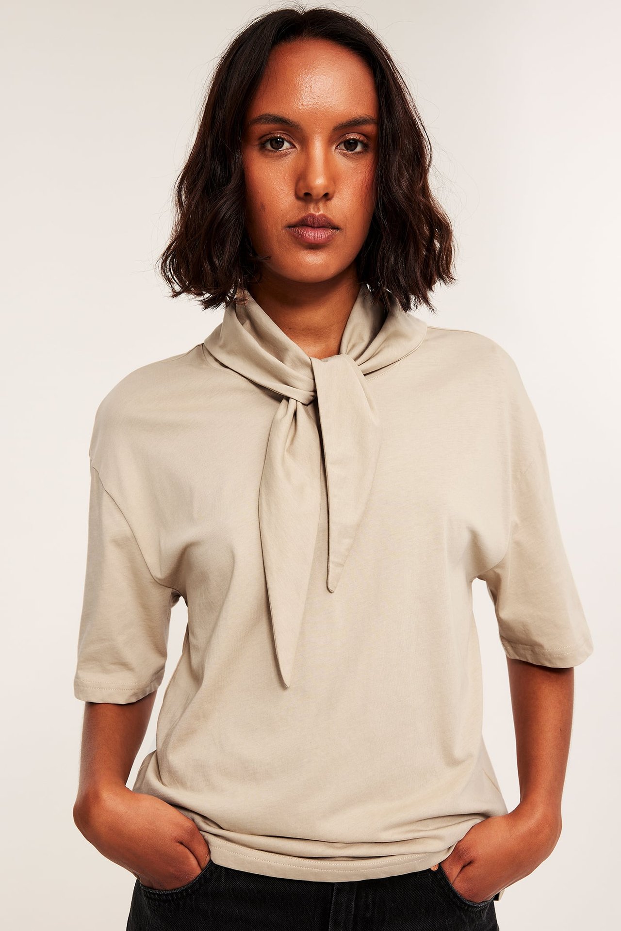 Framsida av kvinnlig modell i ljusbeige t-shirt med halsduk, instoppad i svarta jeans.