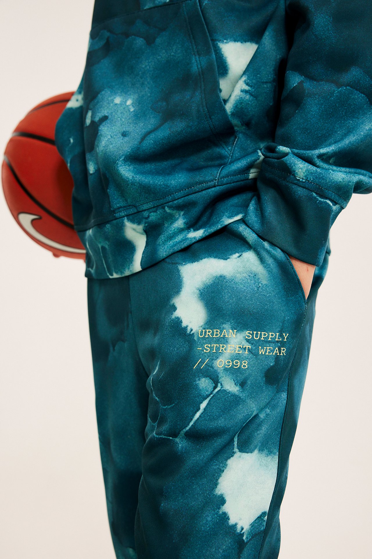 Nærbilde: Mann i turkis tie-dye hettegenser og matchende joggebukse med brodert tekst, holder basketball.