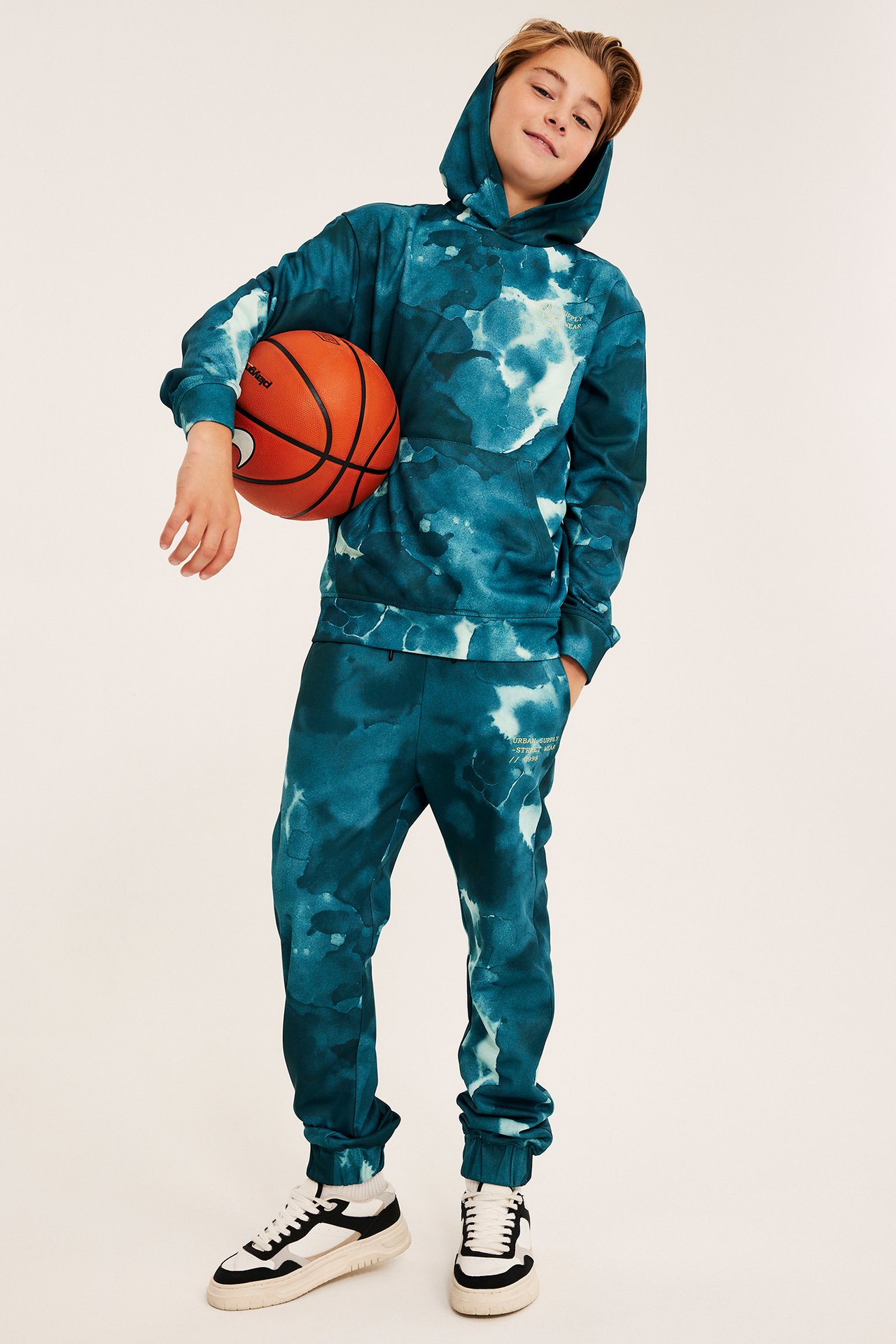 Frontutsikt, et barn i blågrønn tie-dye hettegenser, matchende joggebukse, joggesko, holder en oransje basketball.