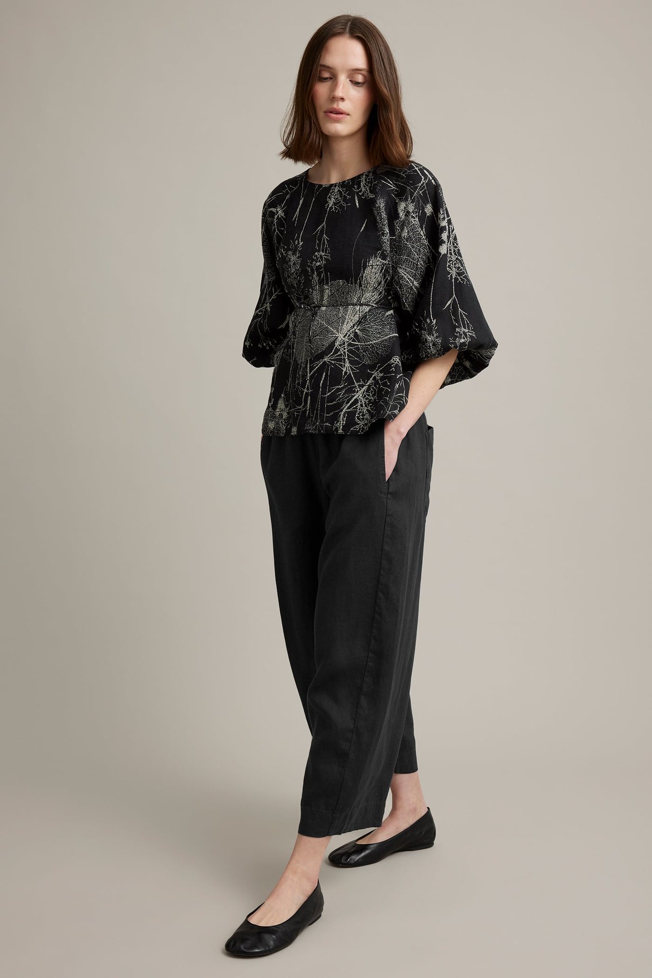 Frontvy av en kvinna i svarta pull-on linnebyxor med barrel fit, en mönstrad blus och svarta ballerinaskor.