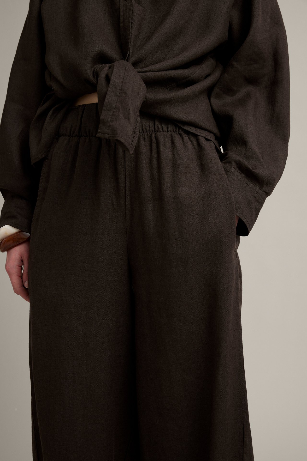 Framifrån: Kvinna i bruna linnebyxor med barrel fit, resår i midjan, knuten skjorta och armband.