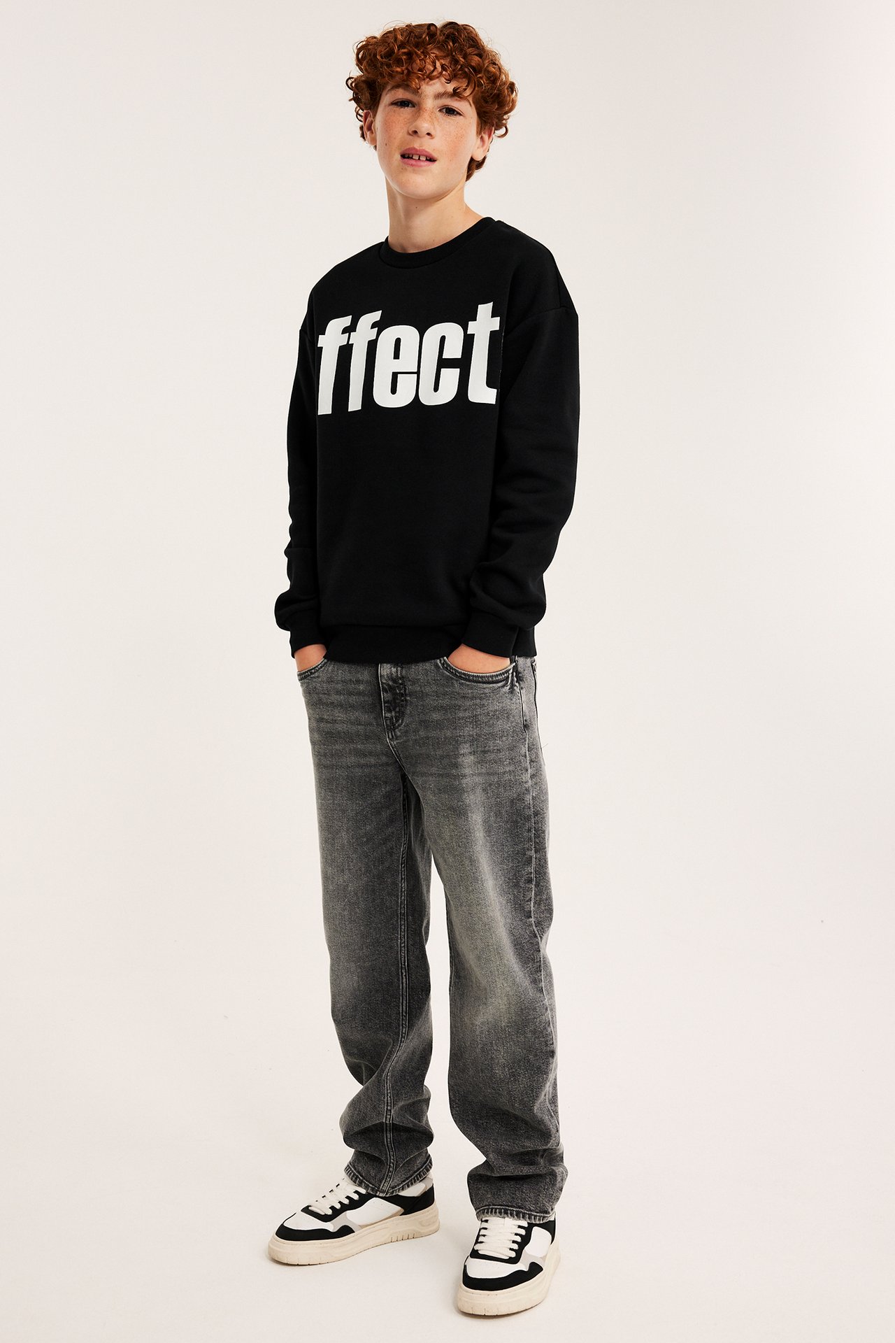 Frontvy av ett barn i svart tröja med 'ffect'-tryck, grå jeans och svartvita sneakers.