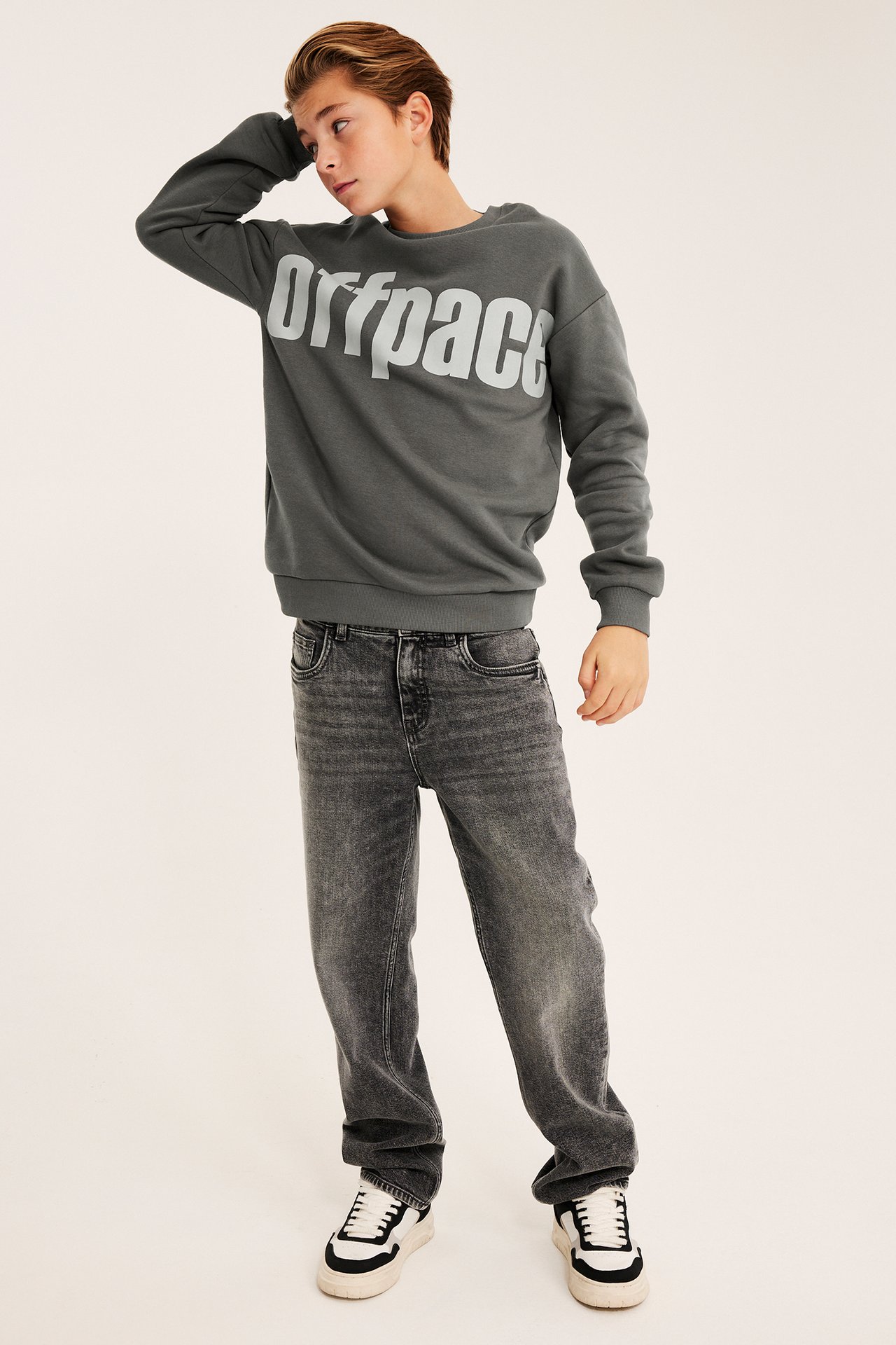 Framsida på barn i mörkgrå sweatshirt med "Offpace"-tryck, tvättade svarta raka jeans och svartvita sneakers.