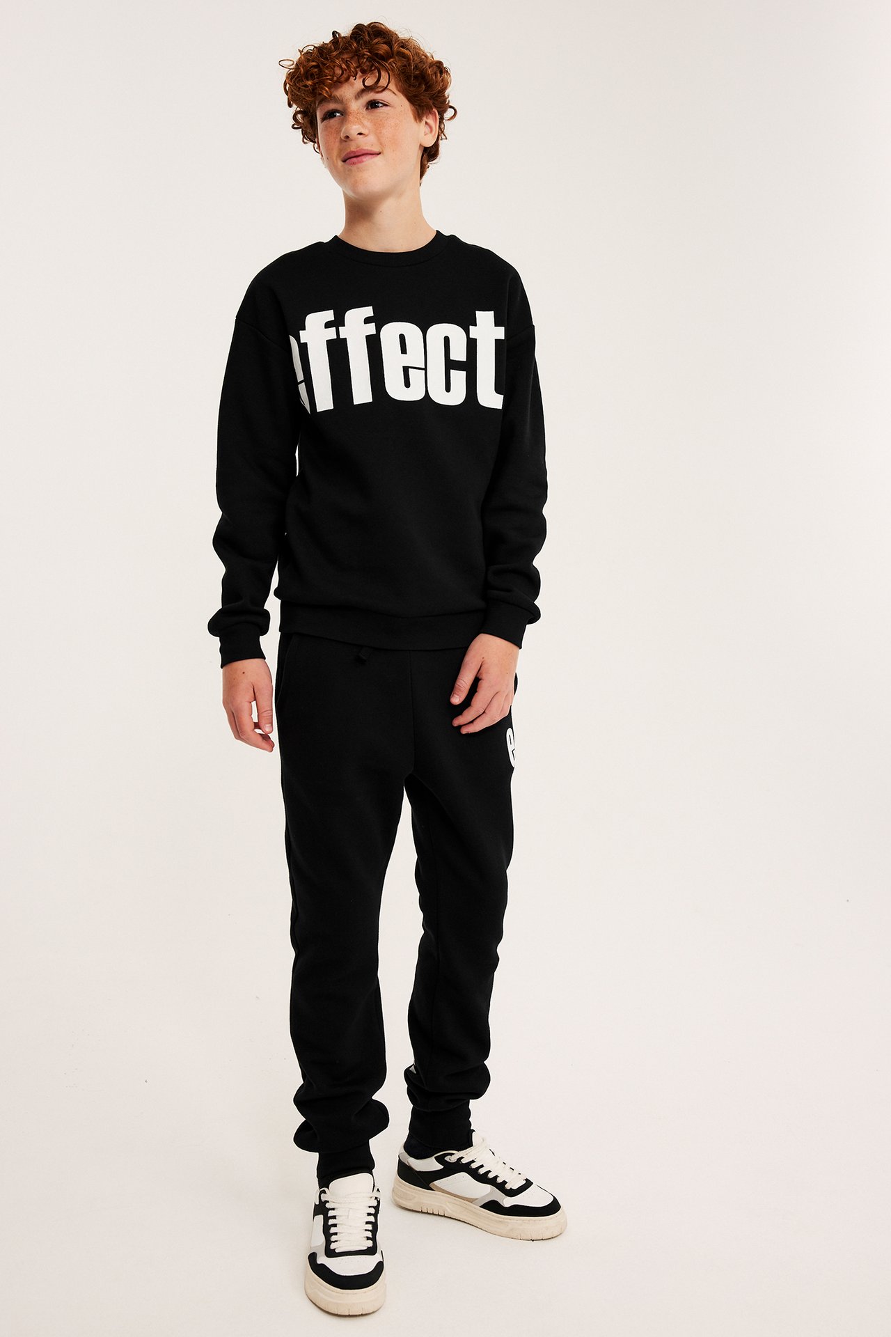 Framifrån, pojke i svart collegetröja med 'fffect' text, svarta mjukisbyxor och svartvita sneakers.