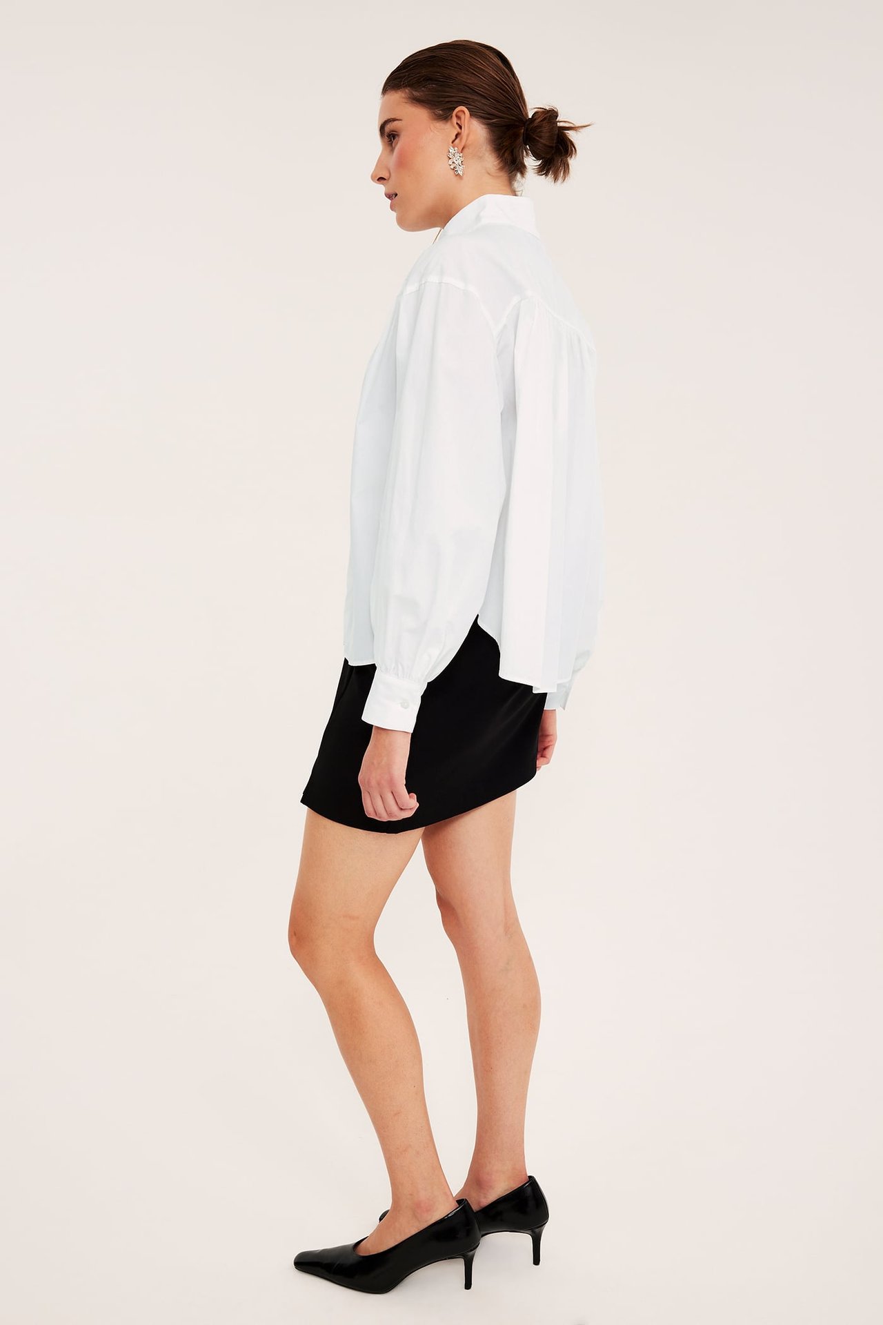 Sidebilde kvinne i hvit oversized skjorte, svart miniskjørt, svarte hæler, sølvøredobber.