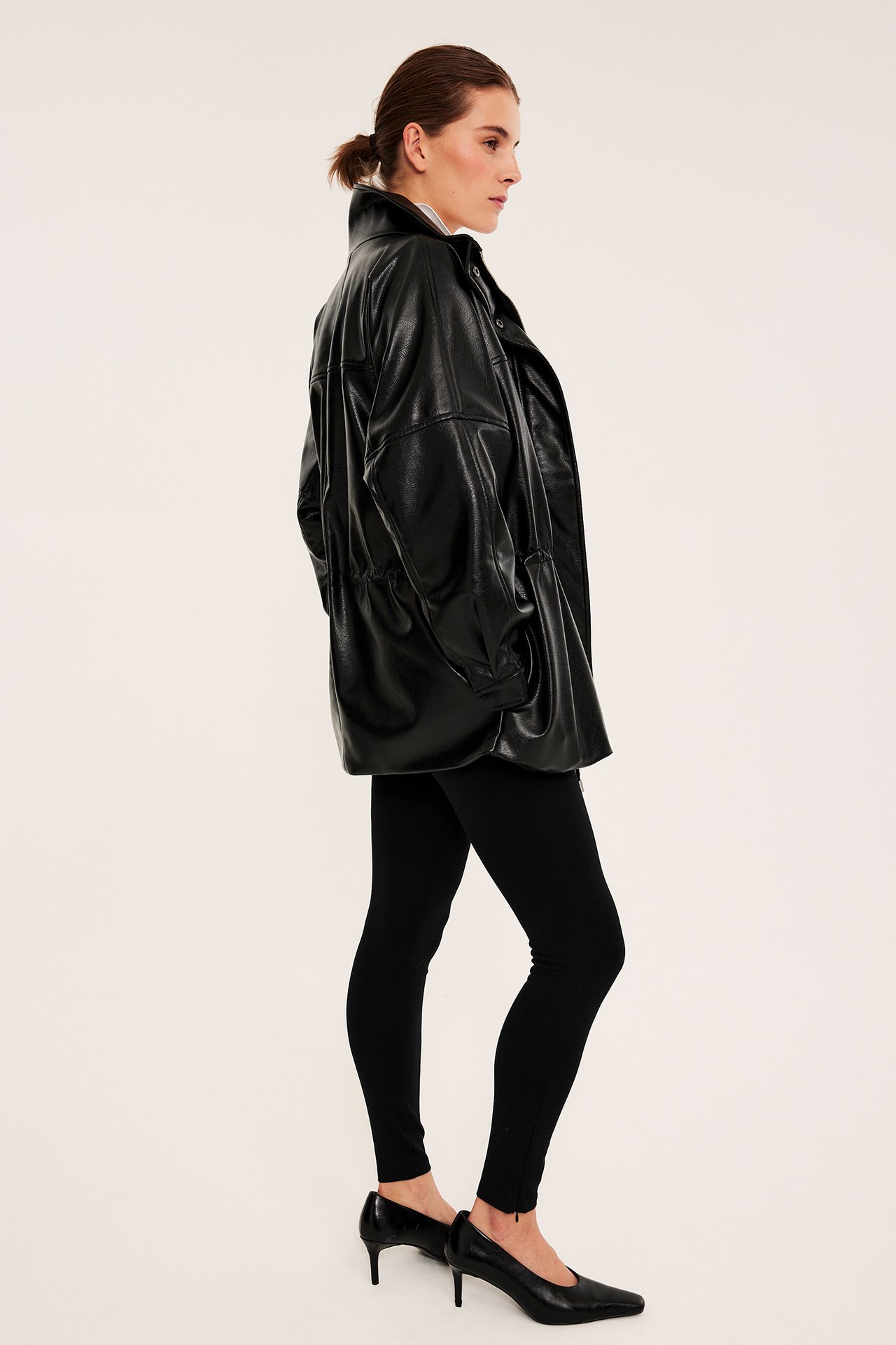 Sidebilde av en kvinne i oversized svart kunstskinnjakke over svarte leggings og svarte pumps.