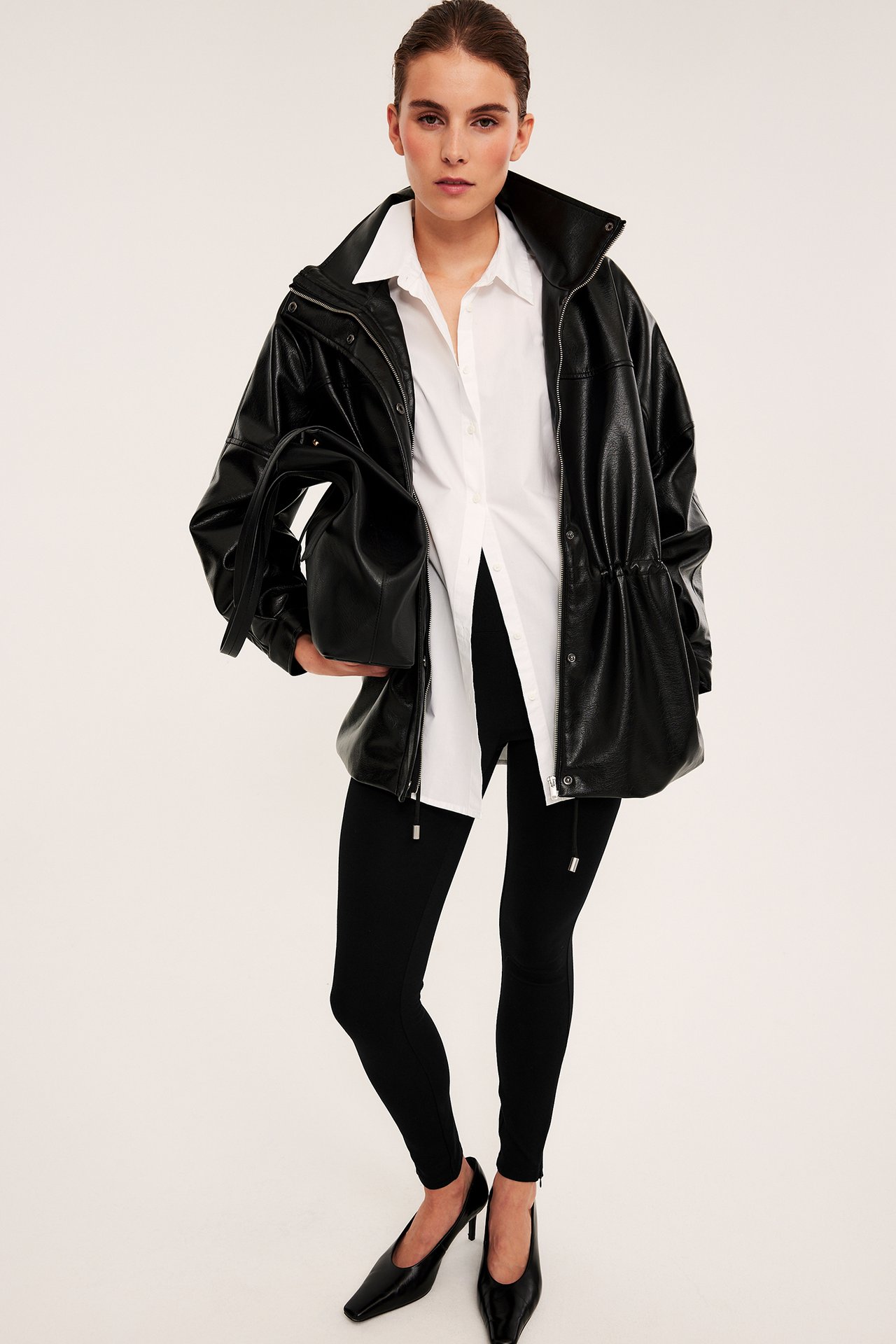 Framsida kvinnlig modell i svart fuskläderjacka, vit skjorta, leggings, klackar, med svart väska.