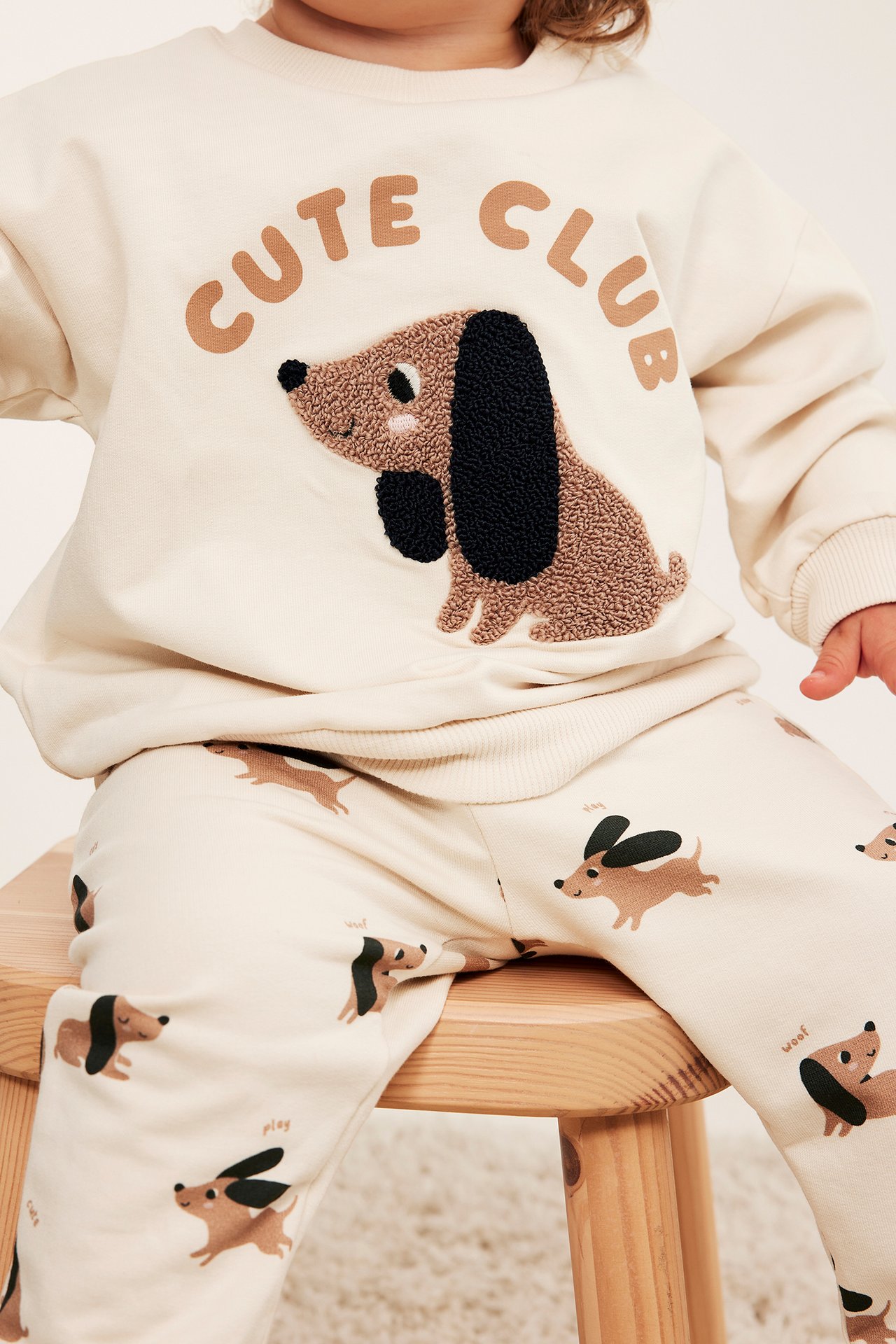 Baby i lys beige sweatshirt med teksturert dachsprint og matchende mønstrede joggebukser.