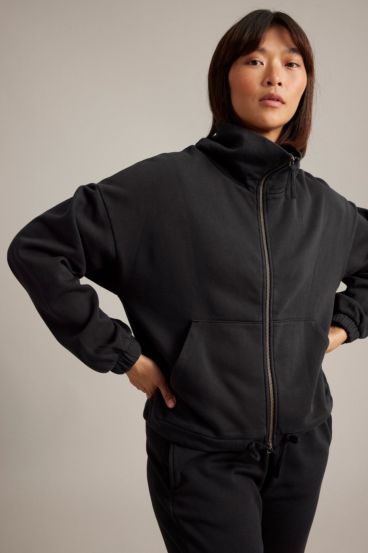 Framifrån en kvinna i svart full-zip sweatshirt med hög krage och svarta sweatpants.