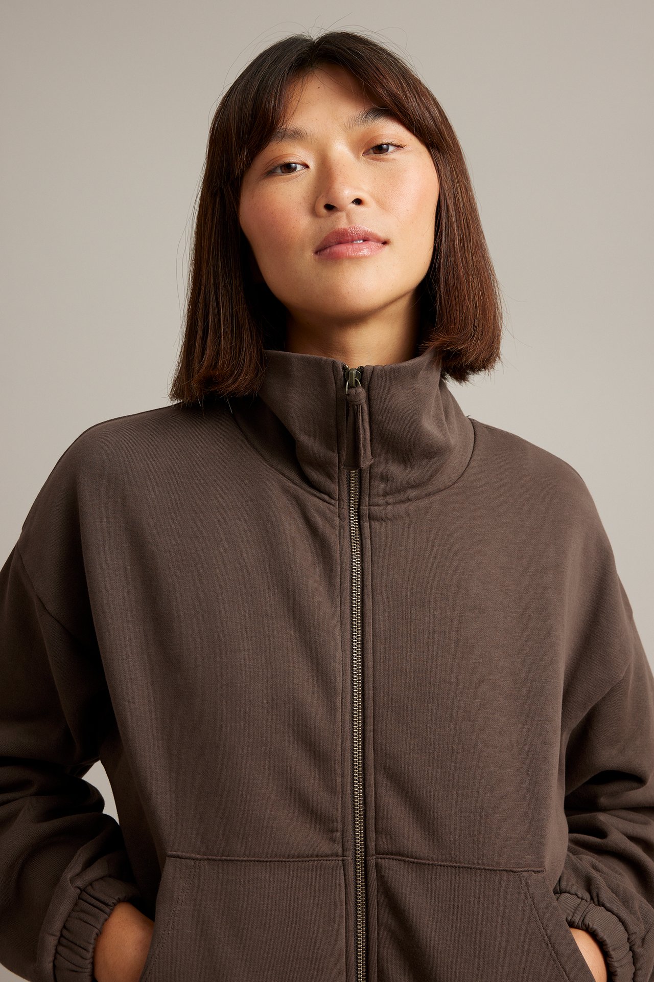 Framsida av en kvinna i mörkbrun hel dragkedja, hög krage sweatshirt med fickor.