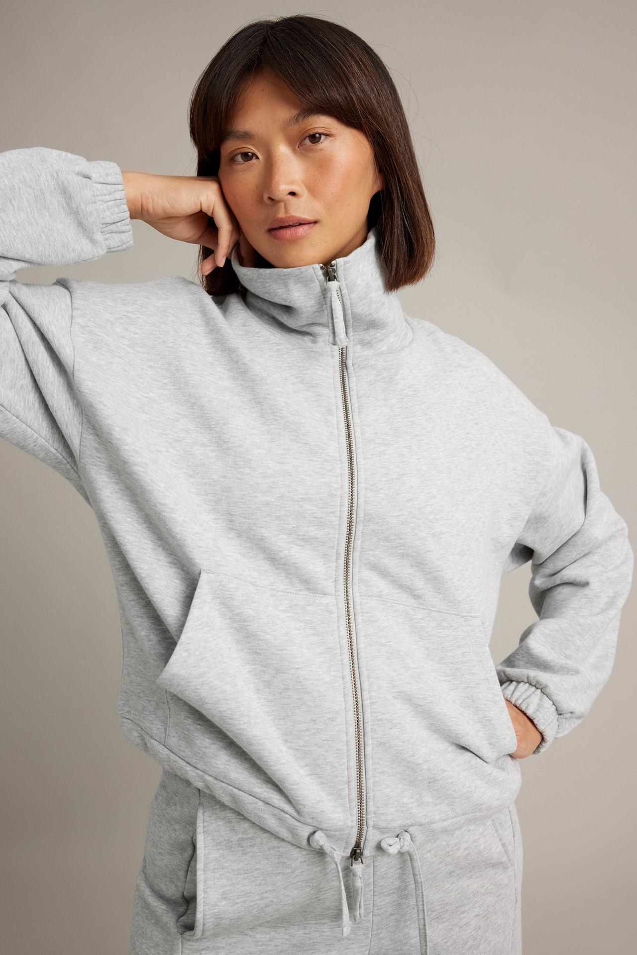 Framsida av kvinna i ljusgrå sweatshirt med dragkedja, fickor och matchande träningsbyxor.