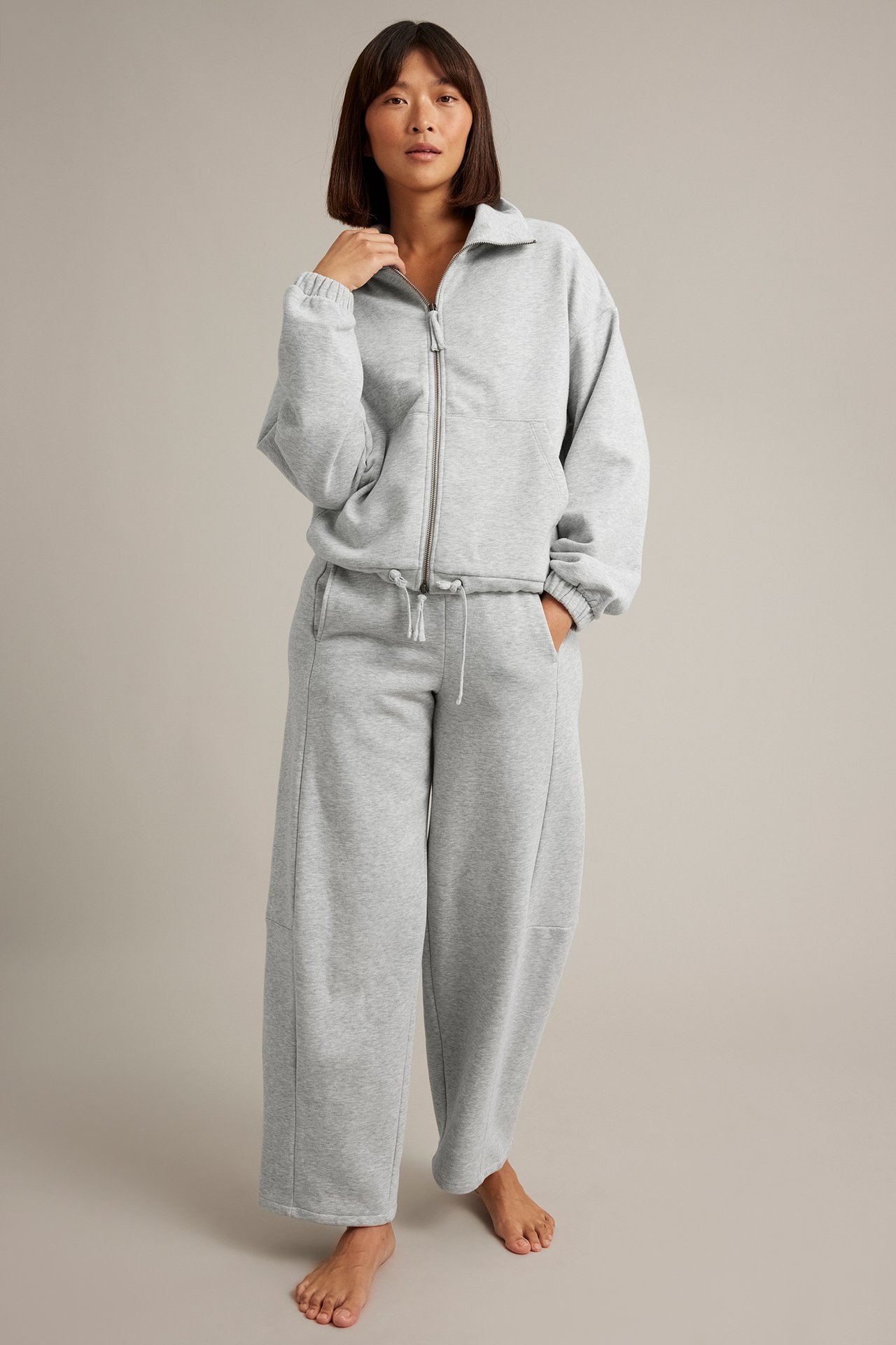 Frontvy av kvinna i gråmelerad sweatshirt med dragkedja och matchande vida sweatpants.