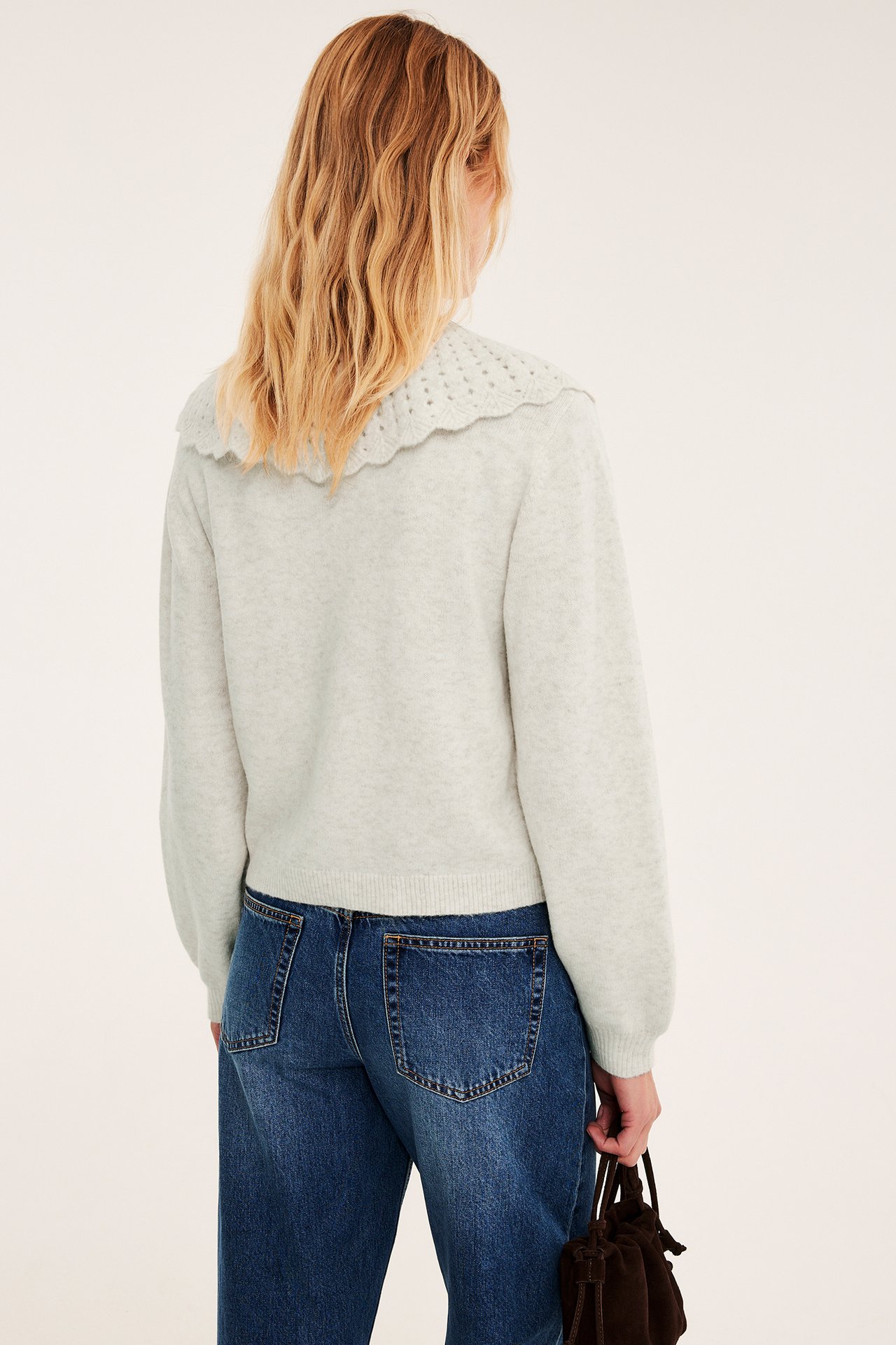 Bakifrån: kvinna i ljusgrå stickad tröja med vågig krage, blå jeans, brun mockaväska.