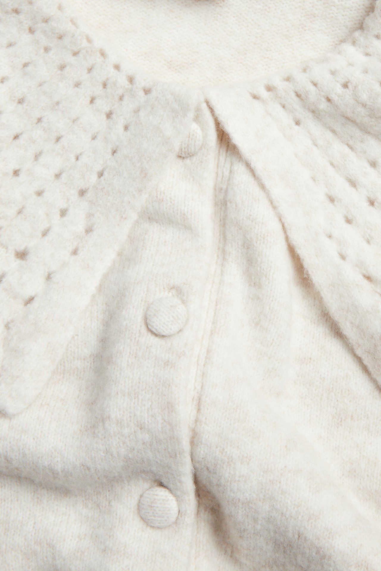 Nærbilde av en lys beige strikket cardigan med hullmønstret krage og stofftrukne knapper.