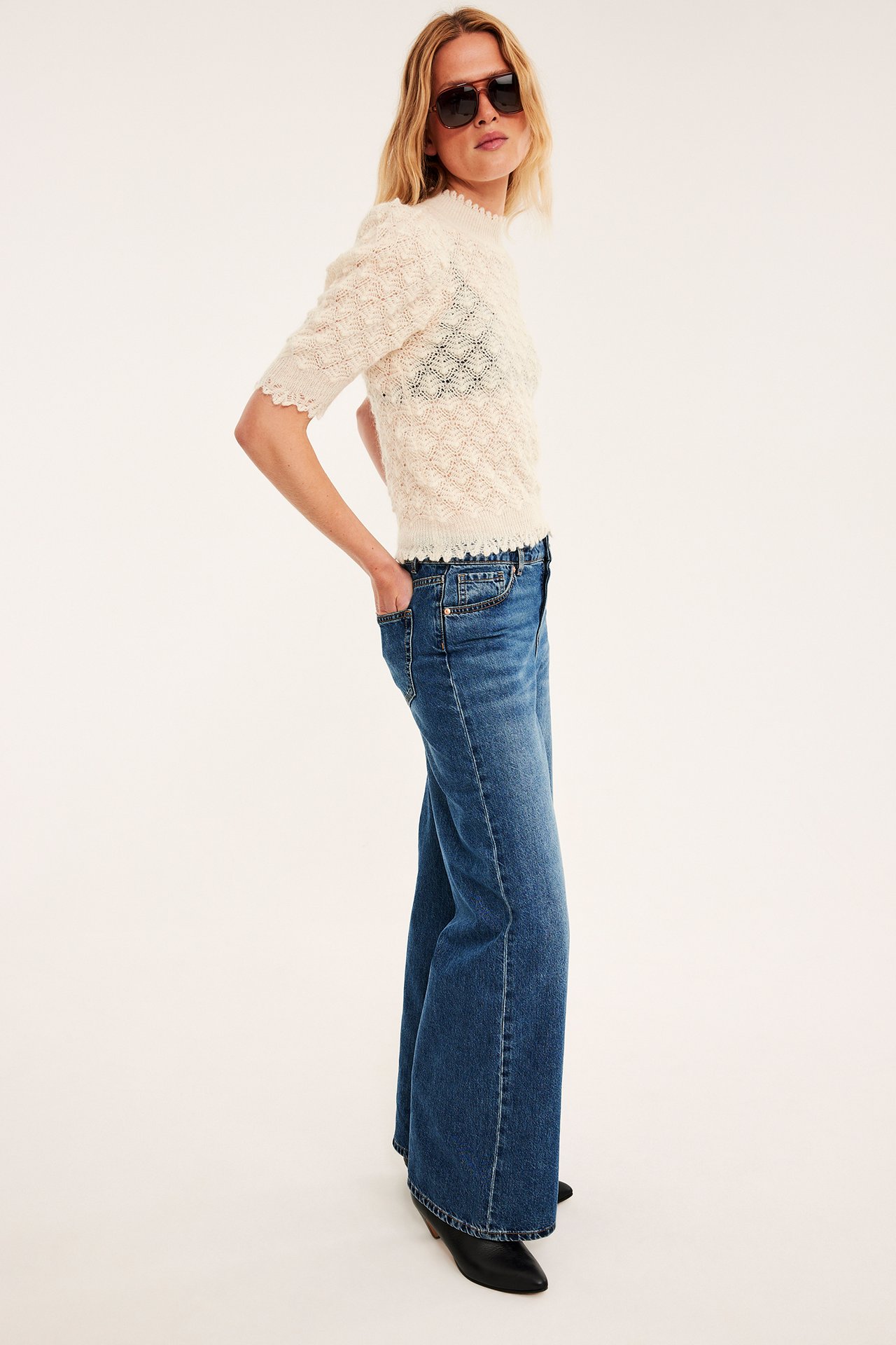 Side, kvinnelig modell i kremfarget åpenstrikket kortermet topp, blå vide jeans, sorte sko.