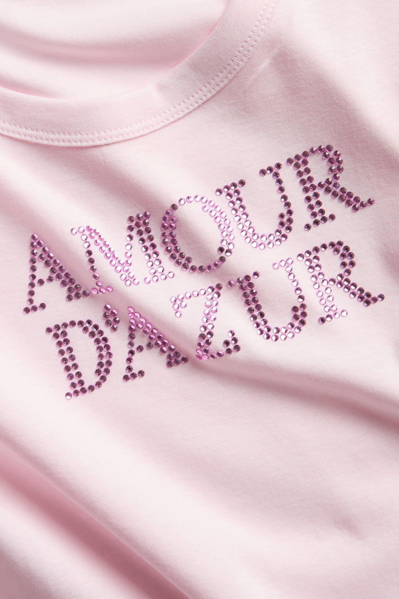 Lähikuva vaaleanpunaisesta t-paidasta, jossa on 'AMOUR D'AZUR' kiiltävillä vaaleanpunaisilla strasseilla.