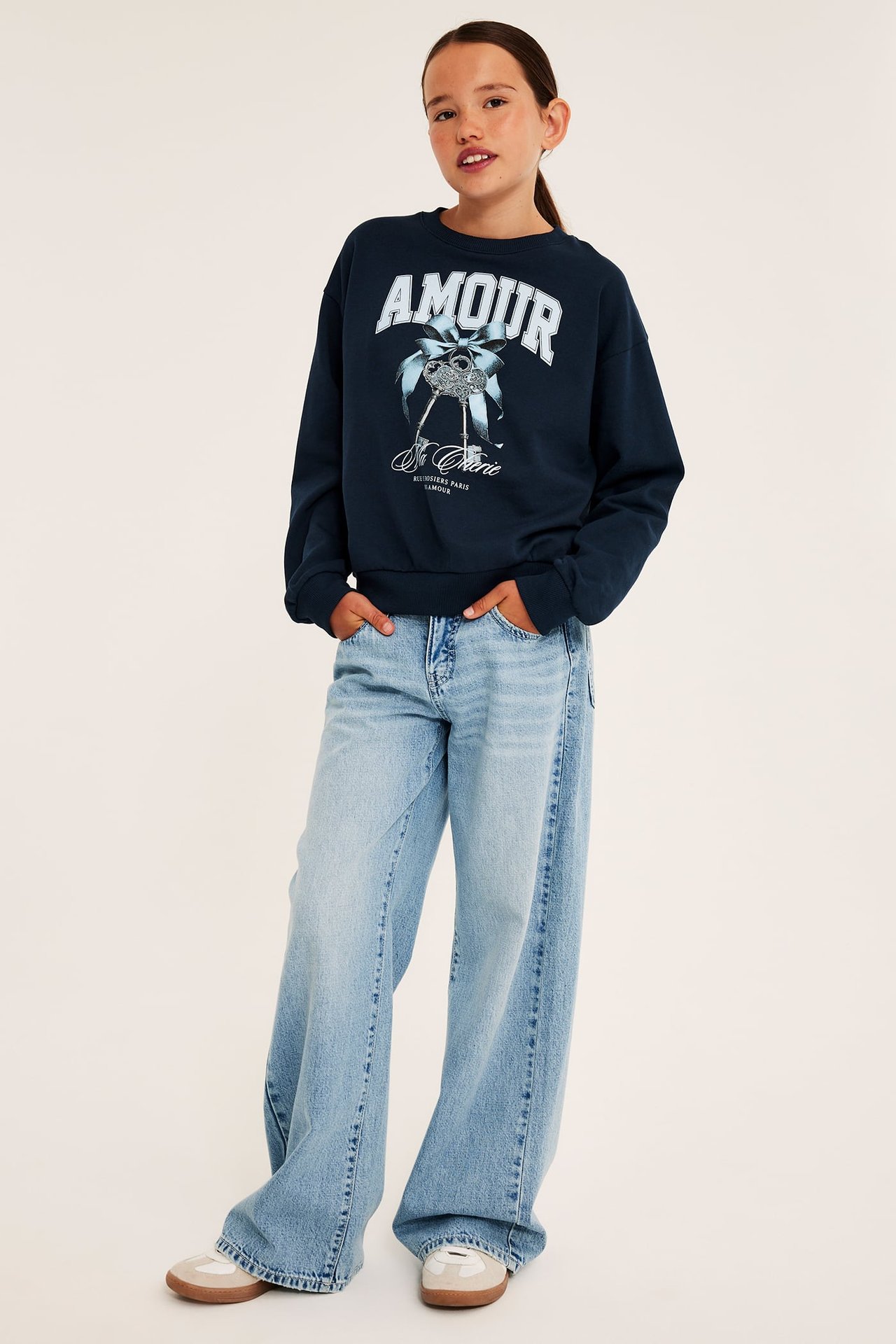 Framifrån: barn i mörkblå 'AMOUR'-tryckt sweatshirt, ljusblå vida jeans och beiga sneakers.