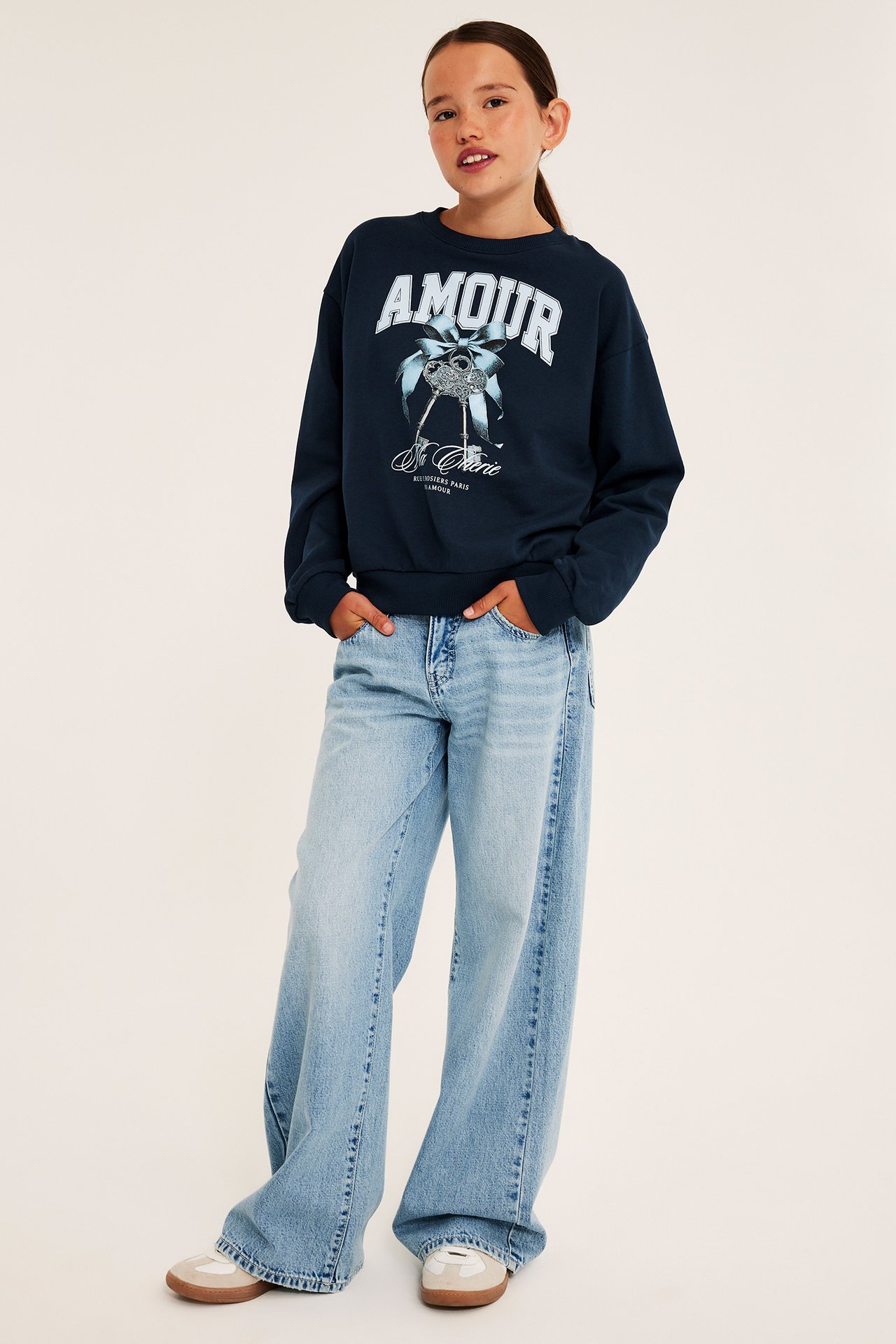Frontbilde av barn i mørkeblå 'AMOUR'-trykt genser, lyseblå vide jeans og beige sneakers.