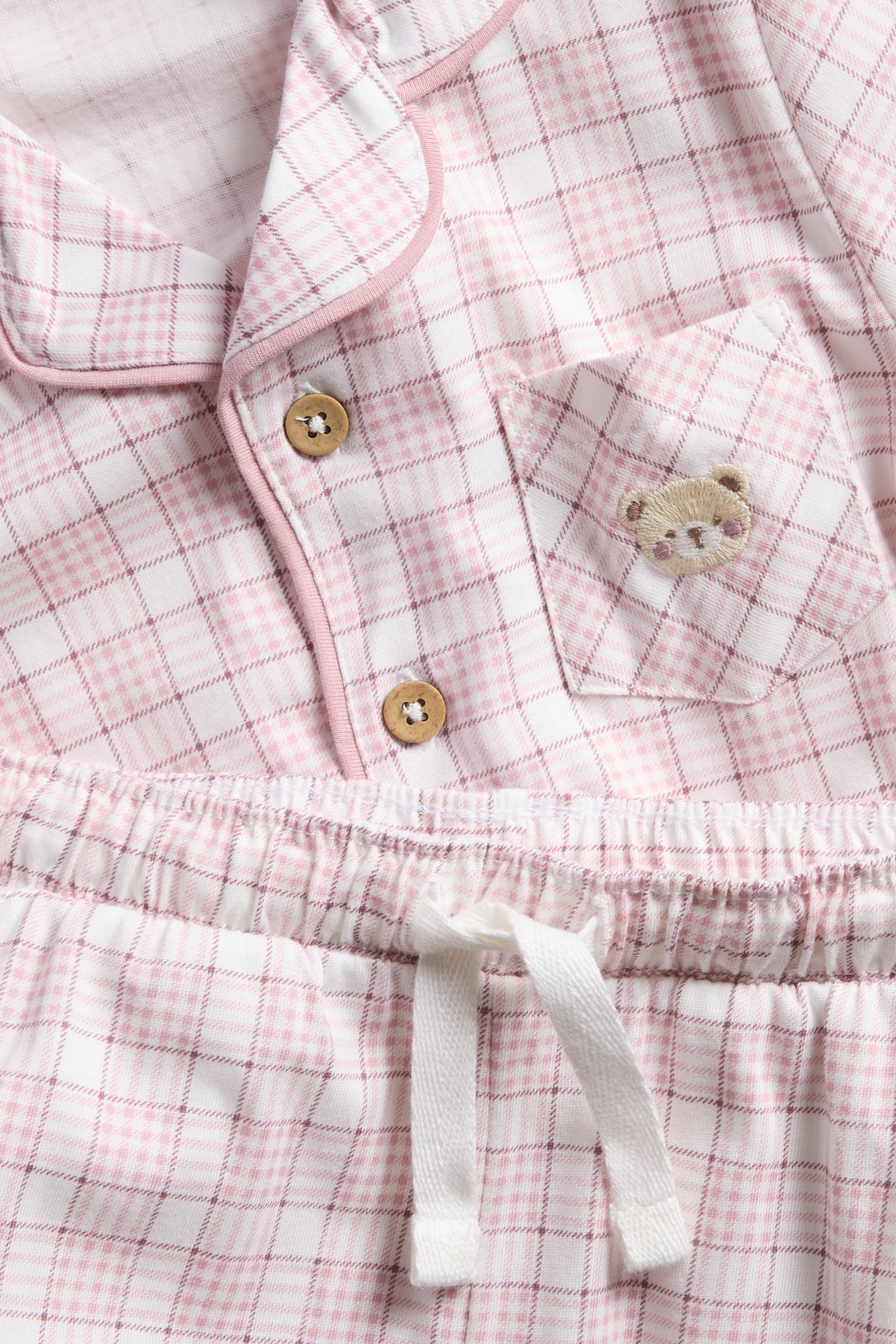 Nærbilde av et rosa og hvitrutete pysjamassett med treknapper og en brodert teddybjørn.