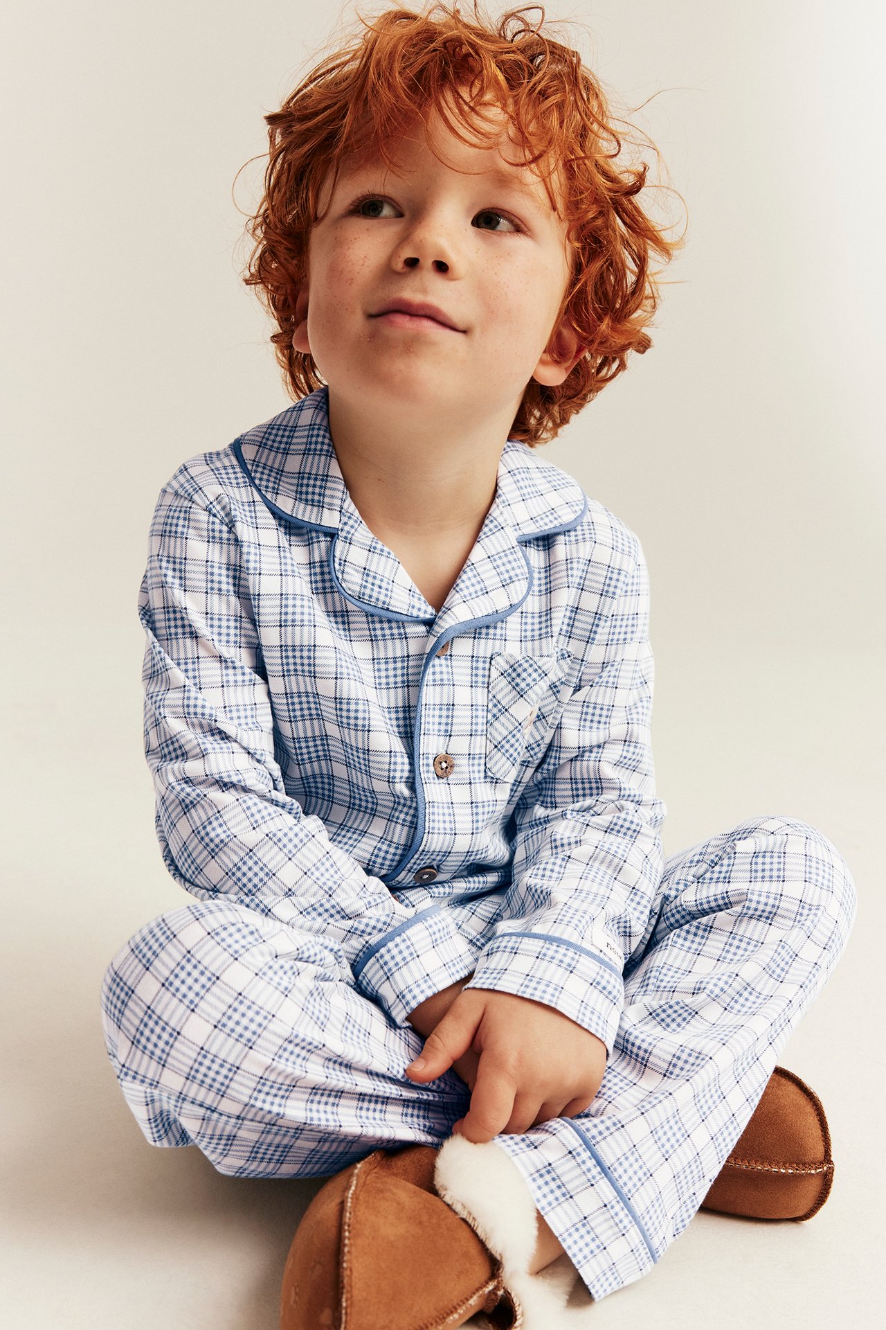 Barn i blå- og hvitrutete pyjamas med knapper og brune tøfler av saueskinn, forfra.