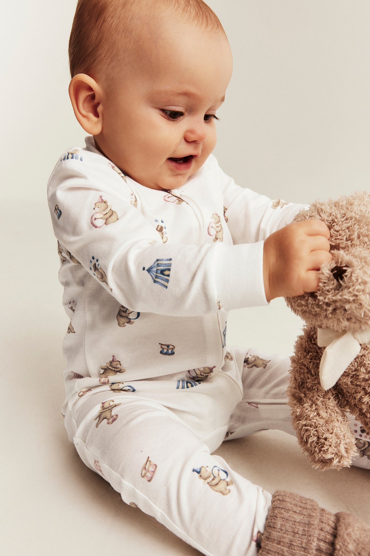 Framifrån, en baby i vit onesie med cirkusdjursmönster och dragkedja håller en brun nallebjörn.
