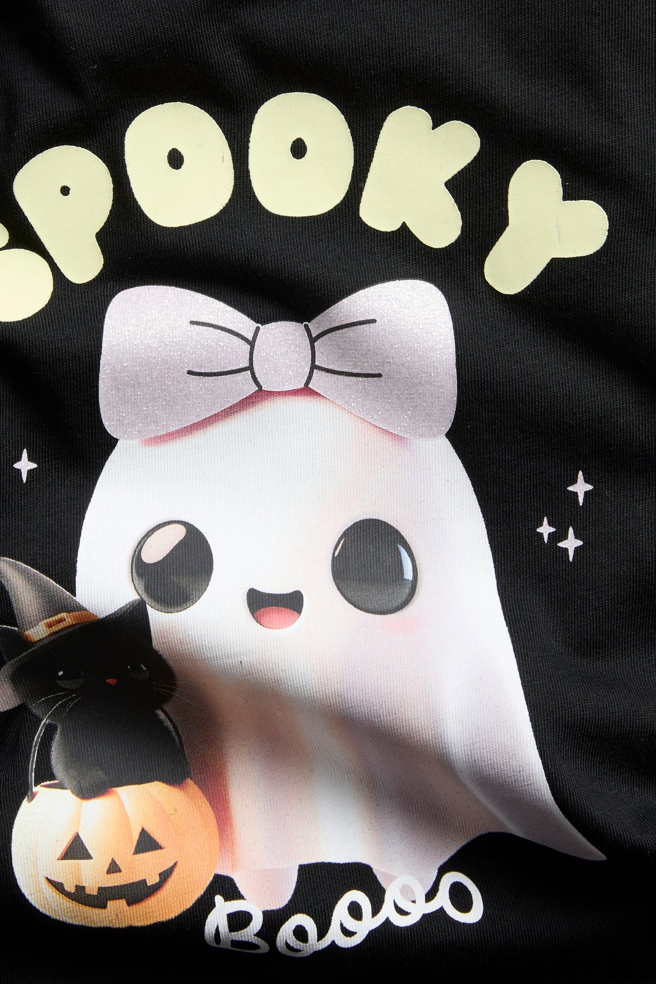 Lähikuva mustasta vaatteesta, jossa söpö Halloween-printti: aave, kissa, kurpitsa ja teksti 'Spooky Booos'.