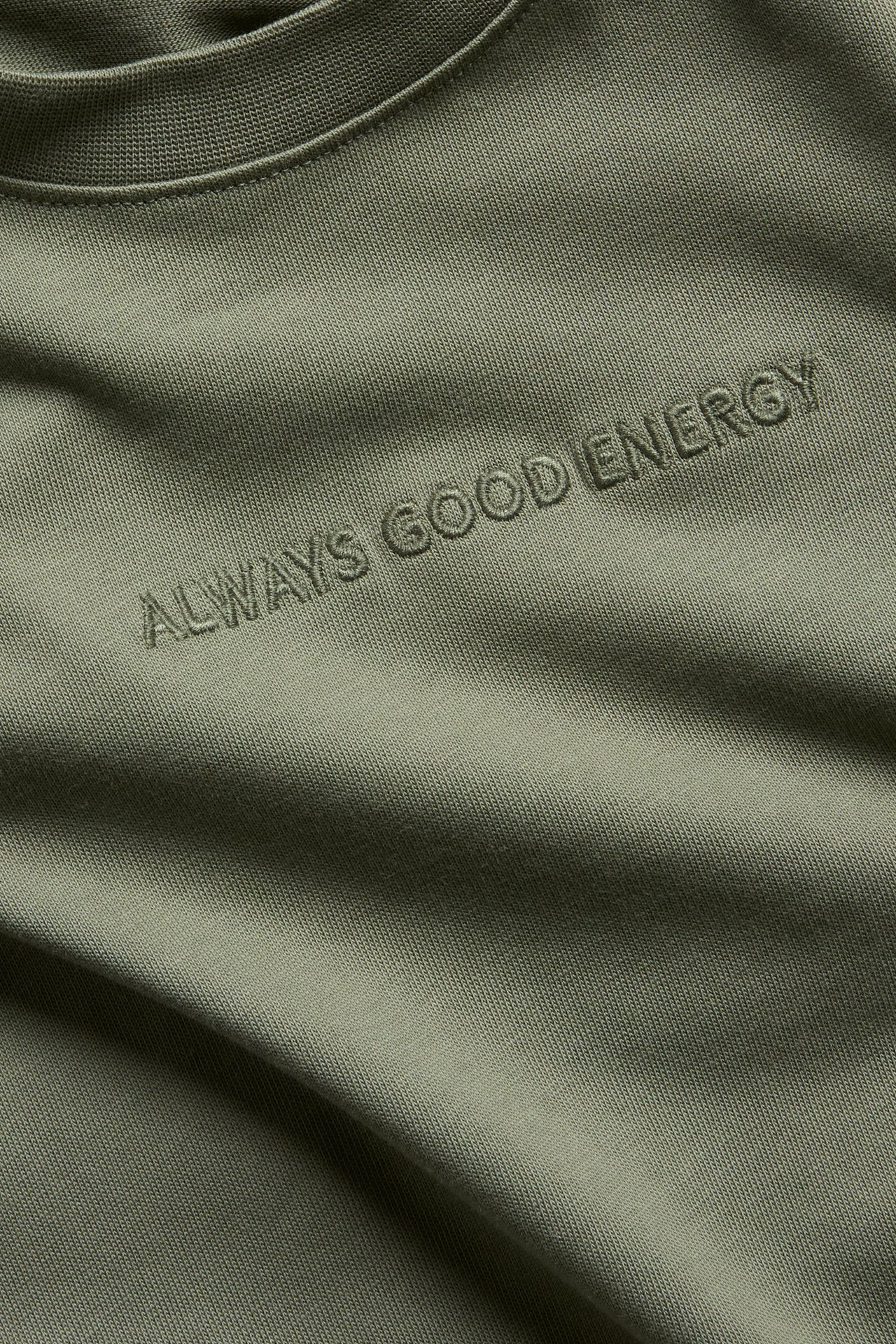 Lähikuva vihreästä lyhythihaisesta T-paidasta, jossa on brodeerattu teksti "ALWAYS GOOD ENERGY" rinnassa.