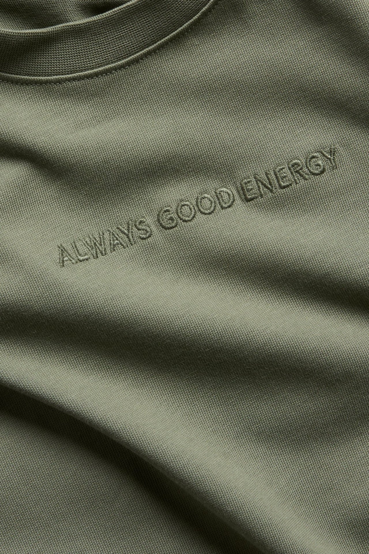 Zbliżenie na zieloną koszulkę z krótkim rękawem z haftowanym napisem „ALWAYS GOOD ENERGY” na piersi.