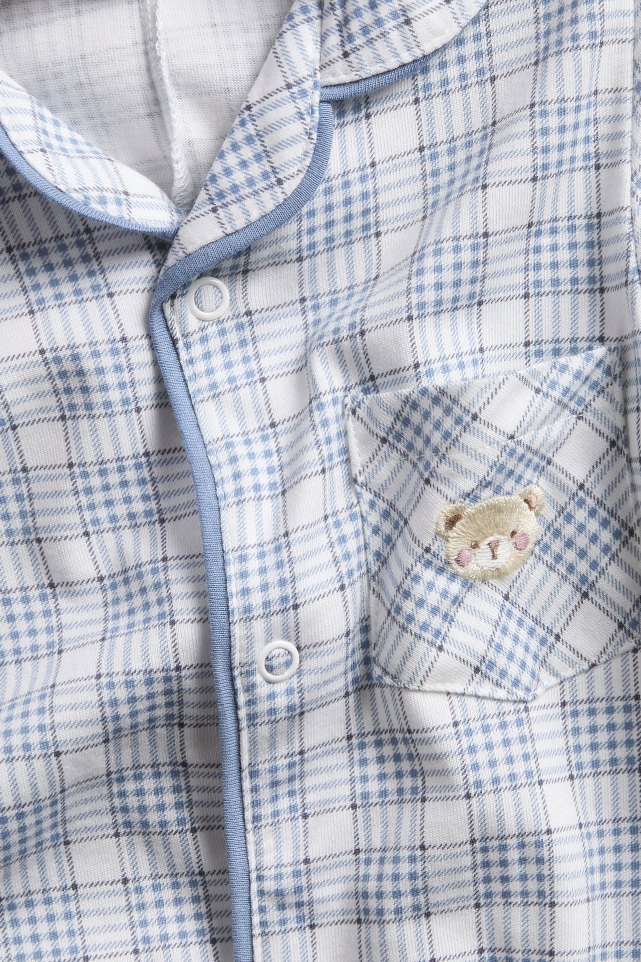 Lähikuva vauvan valko-siniruudullisesta pyjamapaidasta, jossa sininen reunus, nepparit ja brodeerattu karhu.