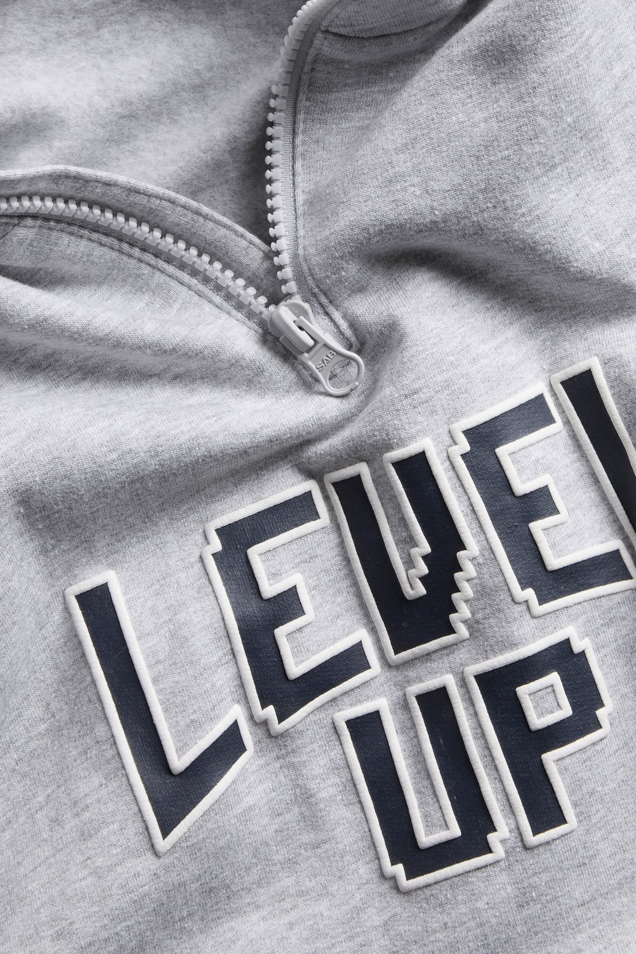 Lähikuva harmaasta collegepaidasta, jossa on muovivetoketju ja pikseloitu ”LEVEL UP” -teksti.