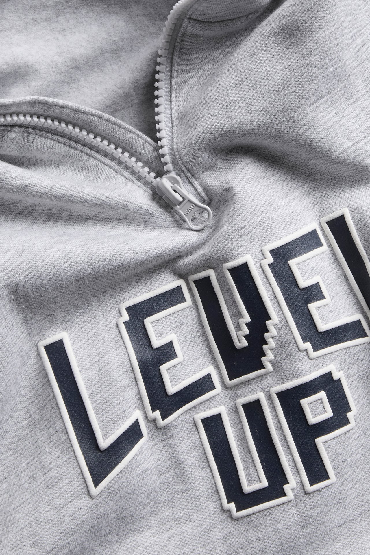 Lähikuva harmaasta collegepaidasta, jossa on muovivetoketju ja pikseloitu ”LEVEL UP” -teksti.