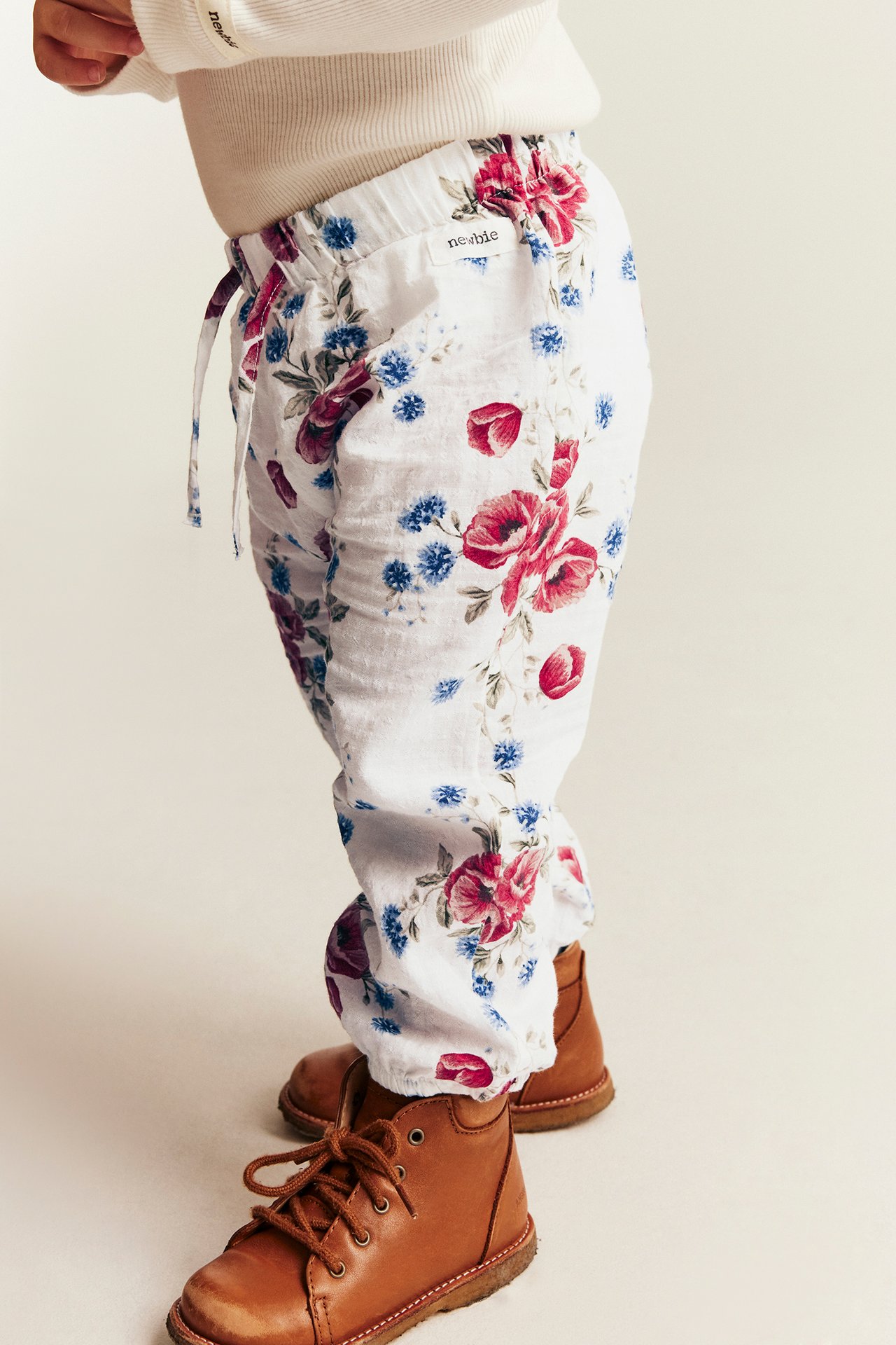 Sidebilde av baby i hvite blomstrete bukser, kremfarget ribbet topp og brune snørestøvler.