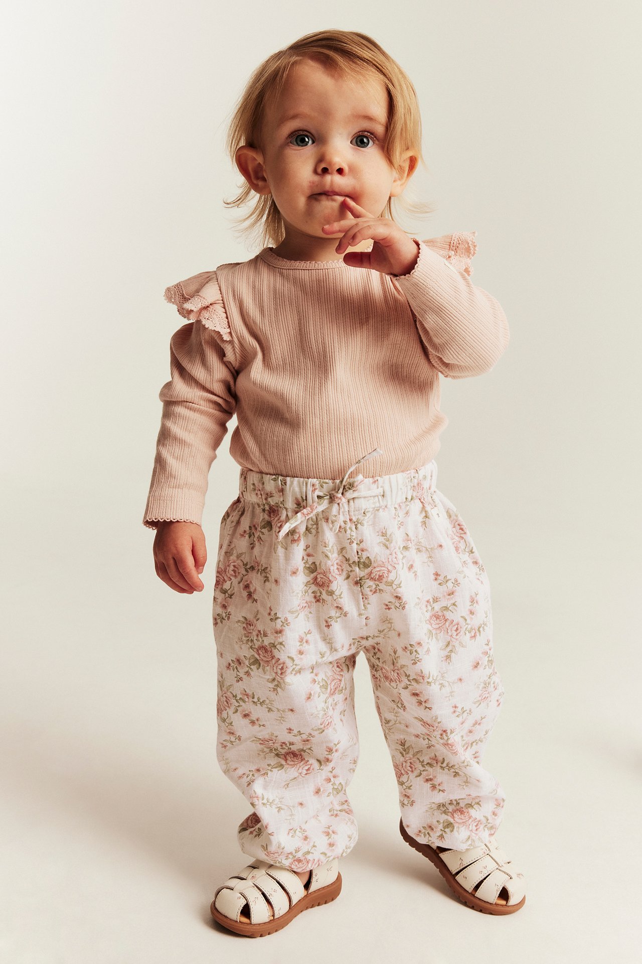 Frontbilde av baby i lys rosa ribbestrikket topp, blomstermønstrede bukser og hvite sandaler.