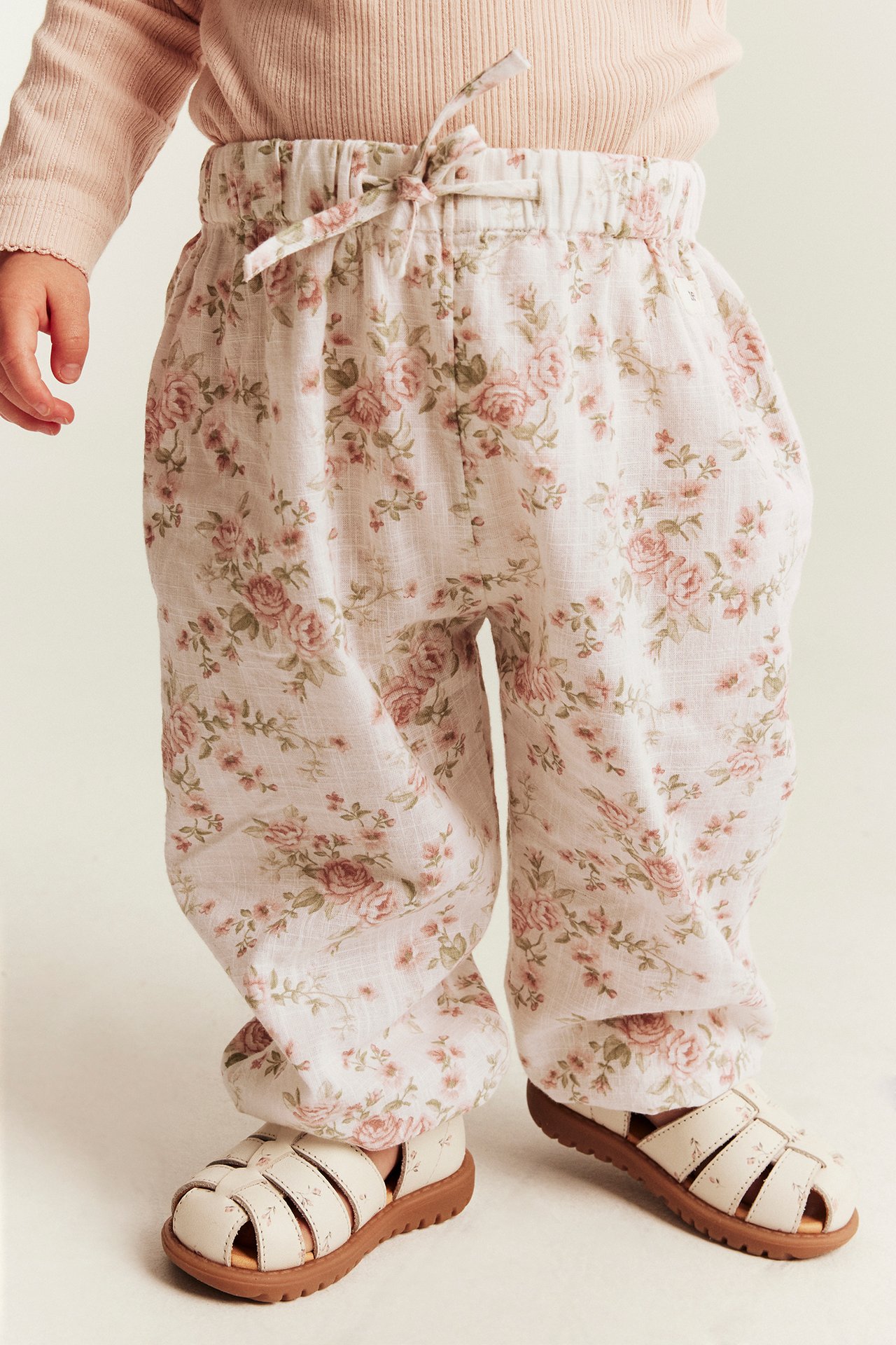 Frontalvisning av baby i rosa blomstrete bukser med knytebånd og kremfargede sandaler.