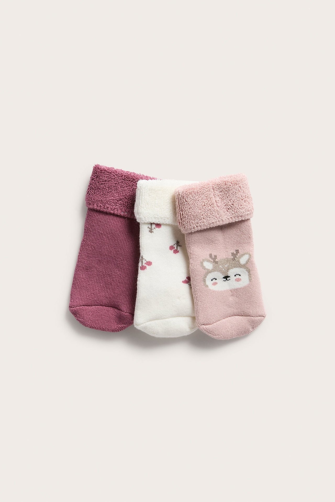 Tre babystrumpor med mudd: enfärgad magenta, vit med körsbär, ljusrosa med rådjursansikte.
