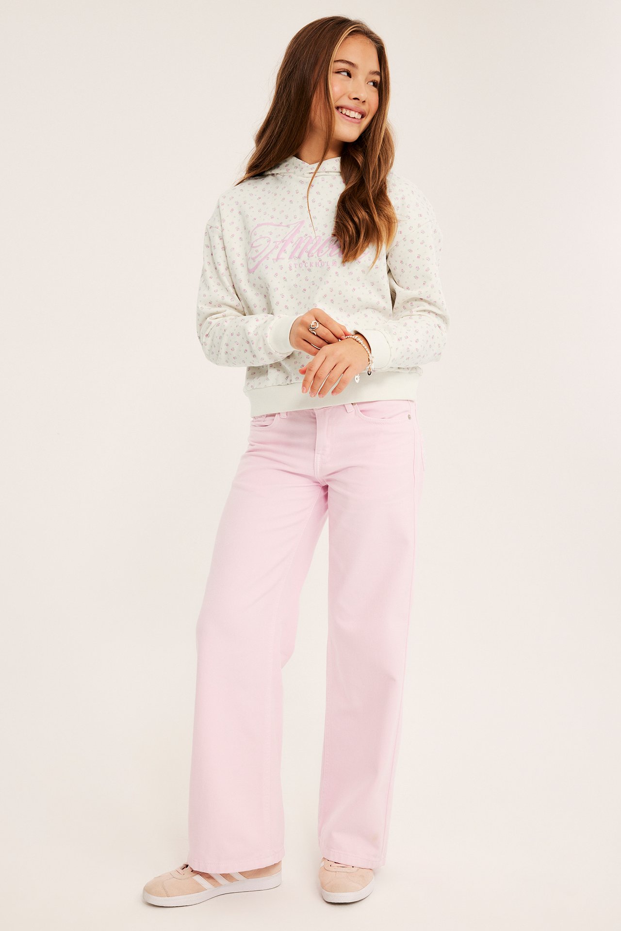 Framsida: Ett barn i vit mönstrad hoodie, rosa vida jeans och ljusrosa sneakers.