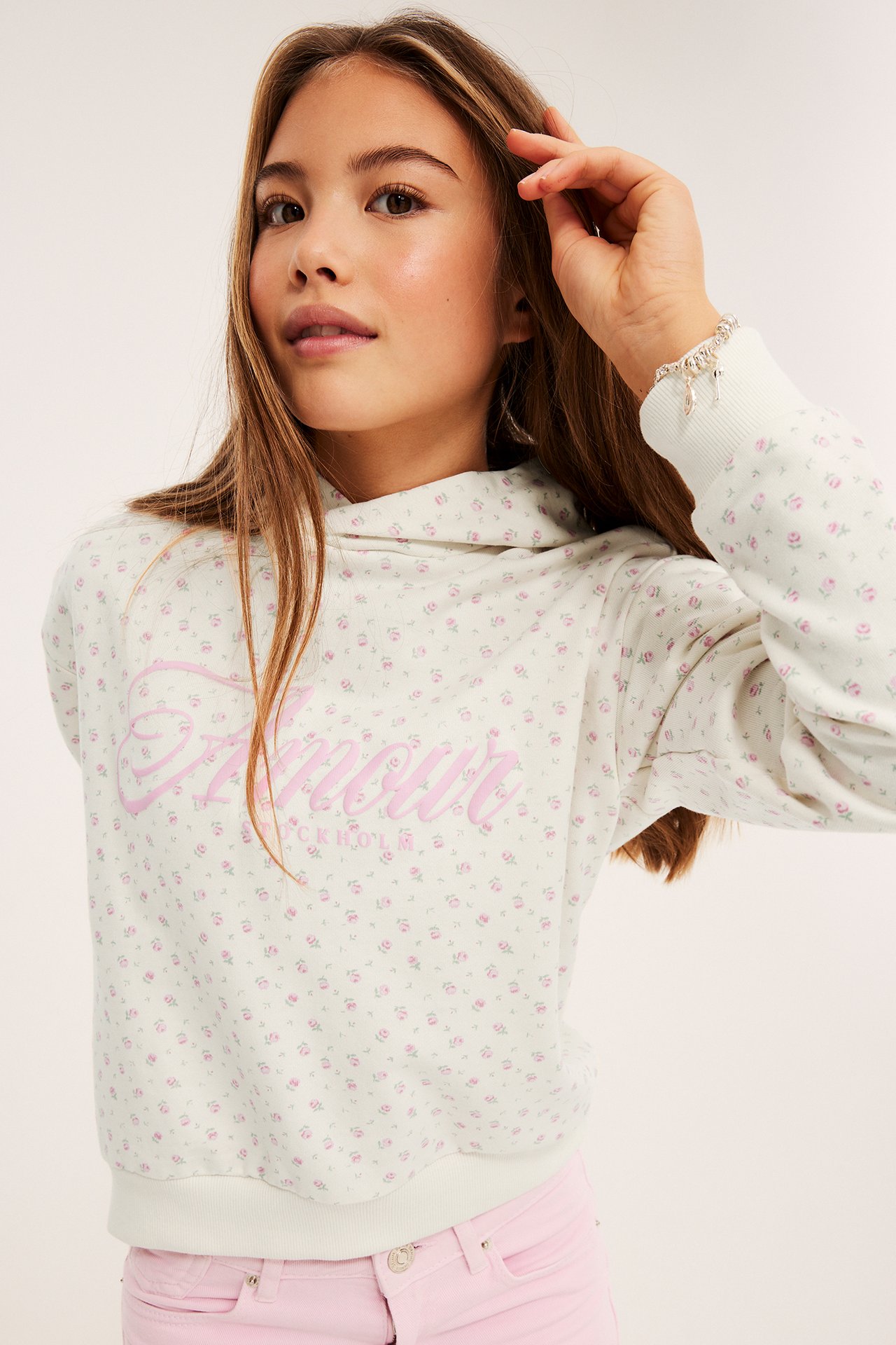 Frontvy, flicka i ljusgrön 'Amour' blommig hoodie, rosa jeans och ett silverarmband med berlocker.