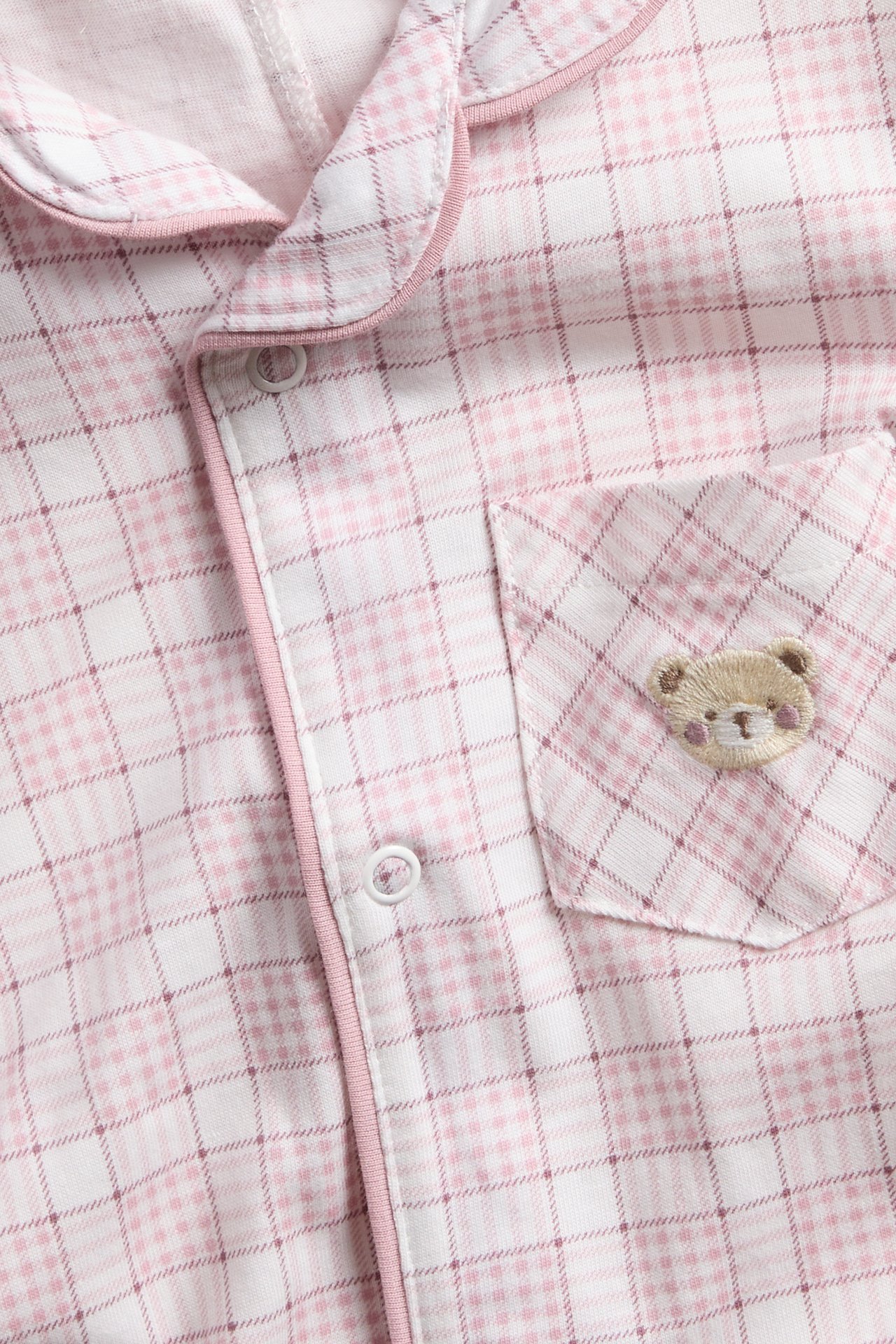 Lähikuva: Pinkki-valkoinen ruudullinen pyjamapaita nallekirjailulla taskussa ja valkoisilla napeilla.
